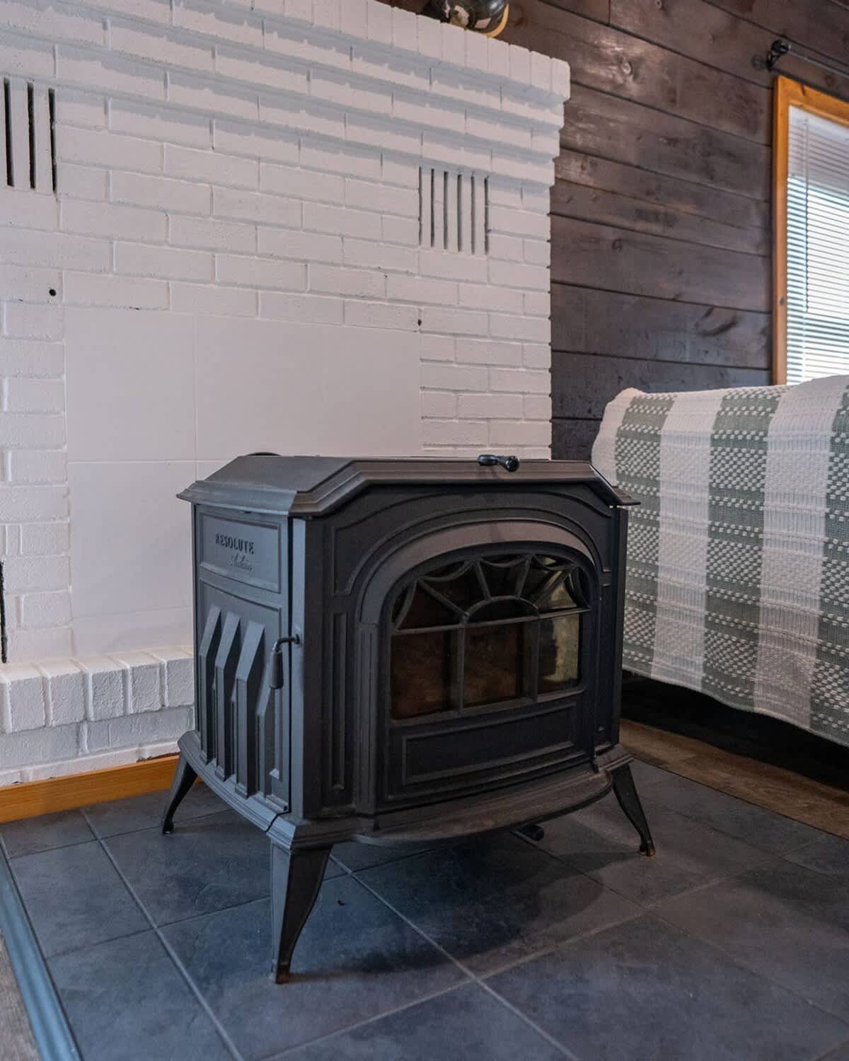 Close-up of a black cast iron wood-burning stove on a dark tiled floor, set against a white brick fireplace and dark wood paneled walls.