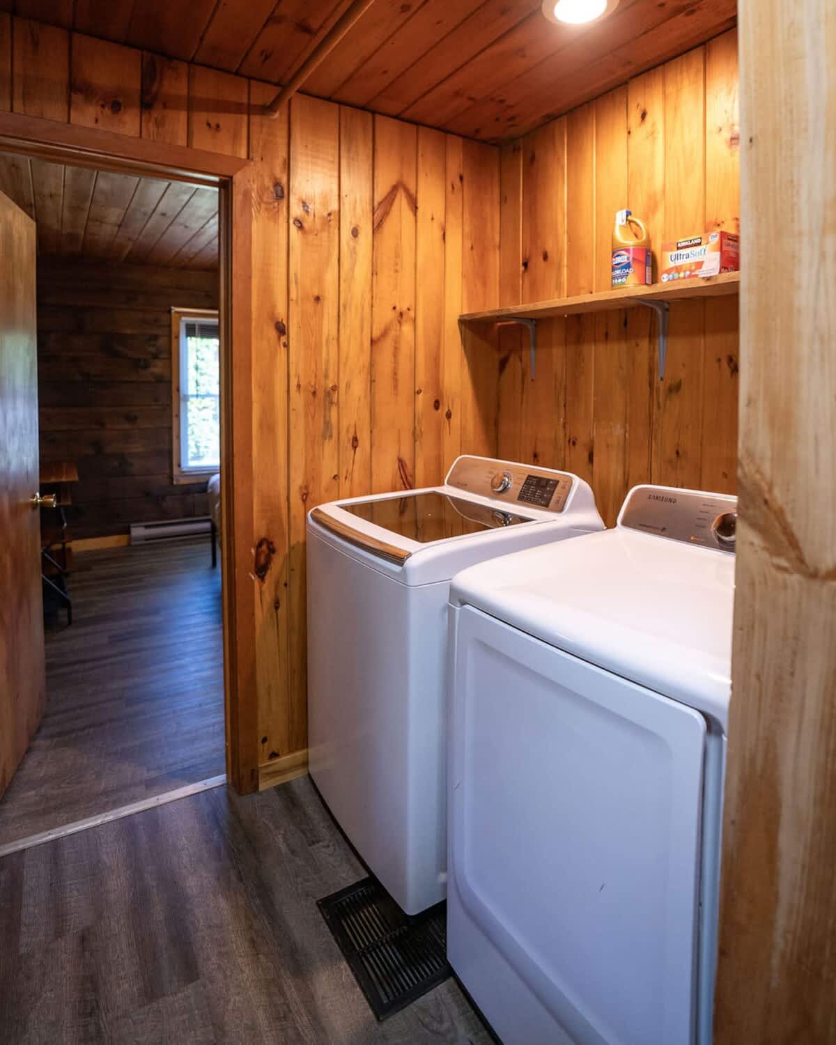 A functional laundry area with warm wood paneled walls and ceiling houses a white top-loading washing machine and a matching front-loading dryer. A simple wooden shelf above holds laundry supplies, and a doorway leads to an adjacent room.