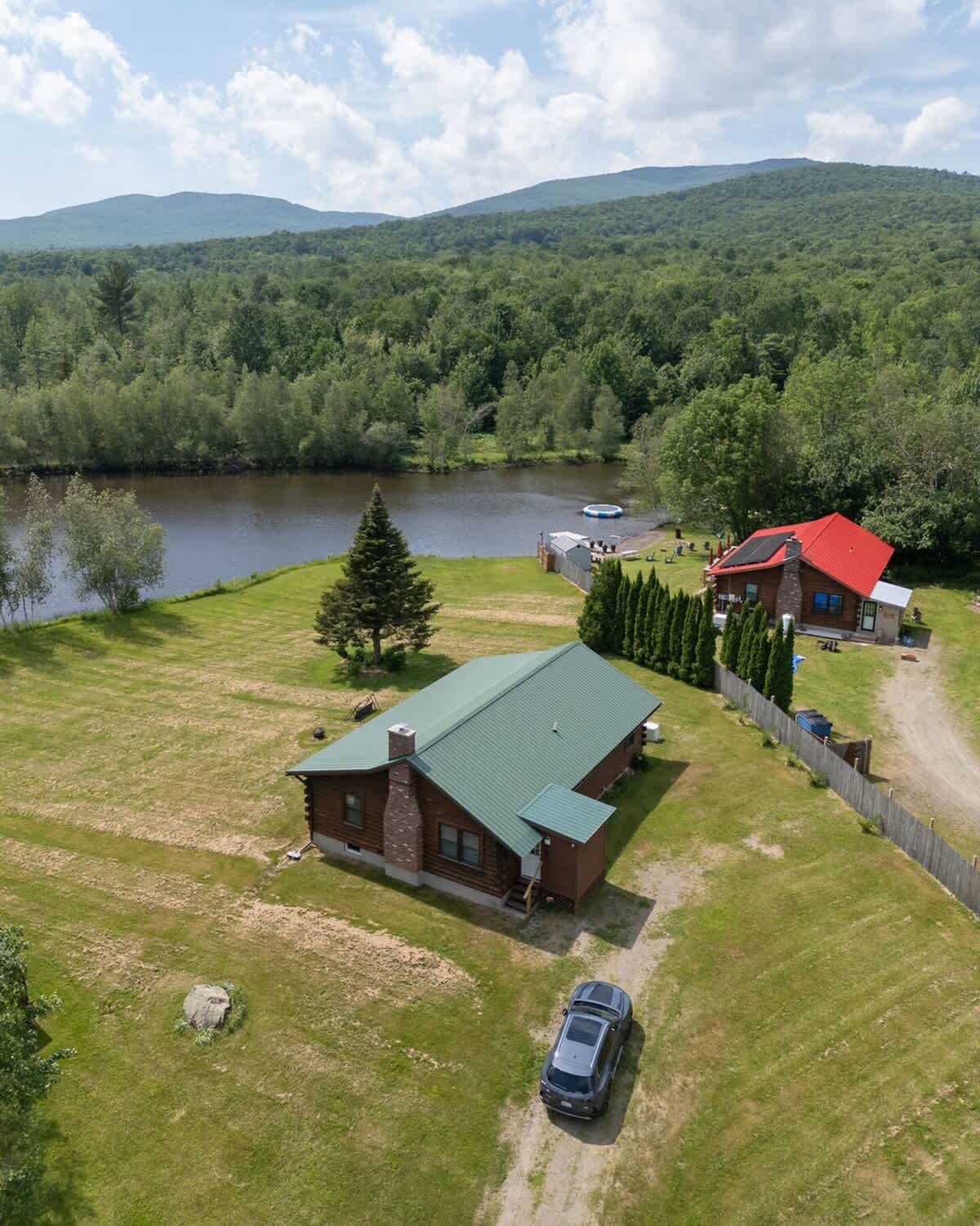 Wide aerial view showcasing the log cabin with a green roof and a second red-roofed building, both situated by a river or lake, surrounded by extensive green landscape and mountains in the distance.