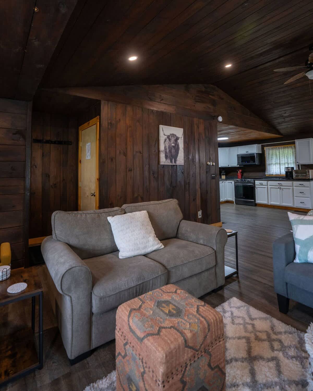 Cozy living room with dark wood paneled walls and ceiling, featuring a gray sofa with a white pillow, a patterned ottoman, and a glimpse of a kitchen in the background.
