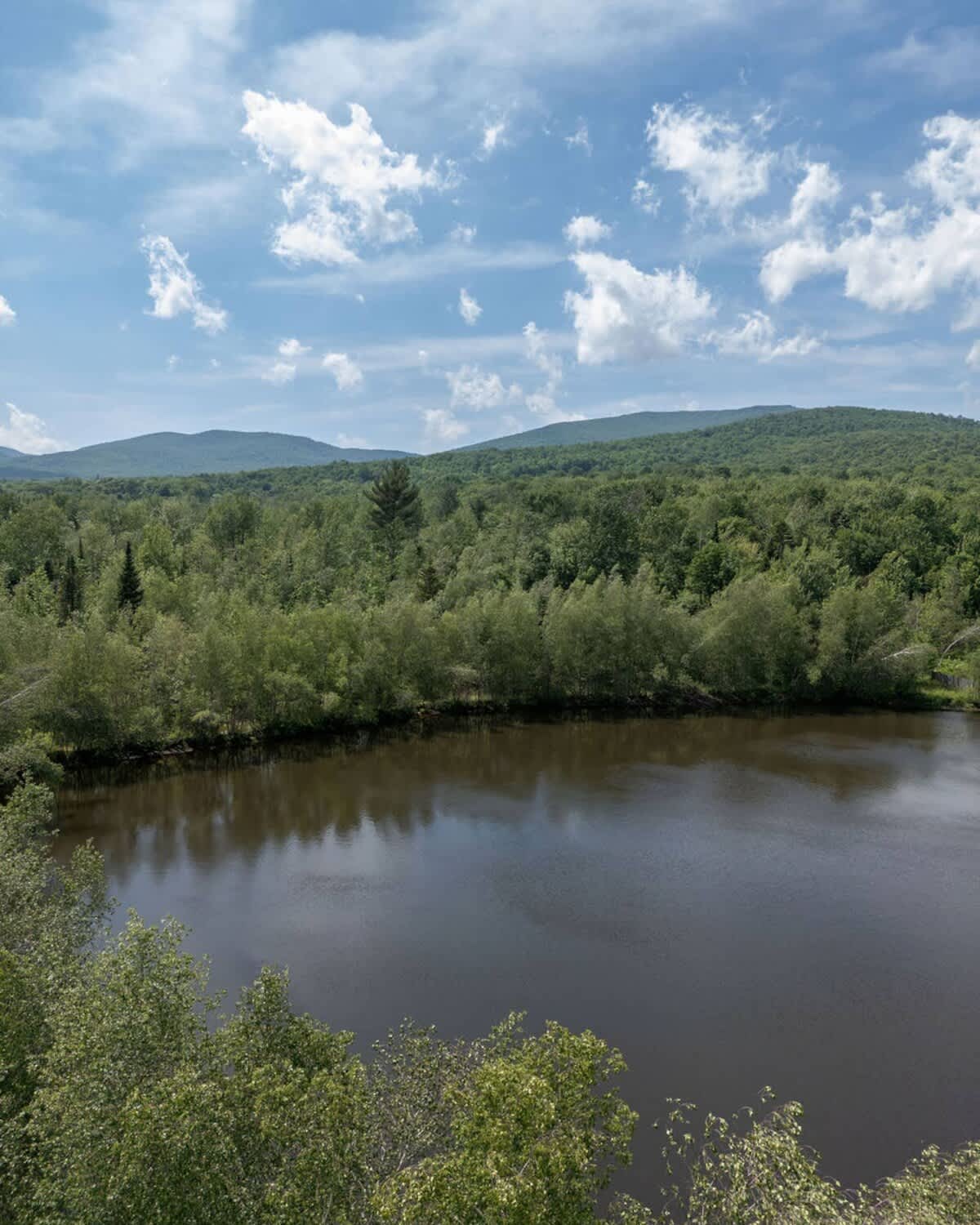 Expansive scenic view of a dark lake surrounded by dense green forests, with rolling mountains visible in the distance under a bright blue sky with fluffy white clouds.