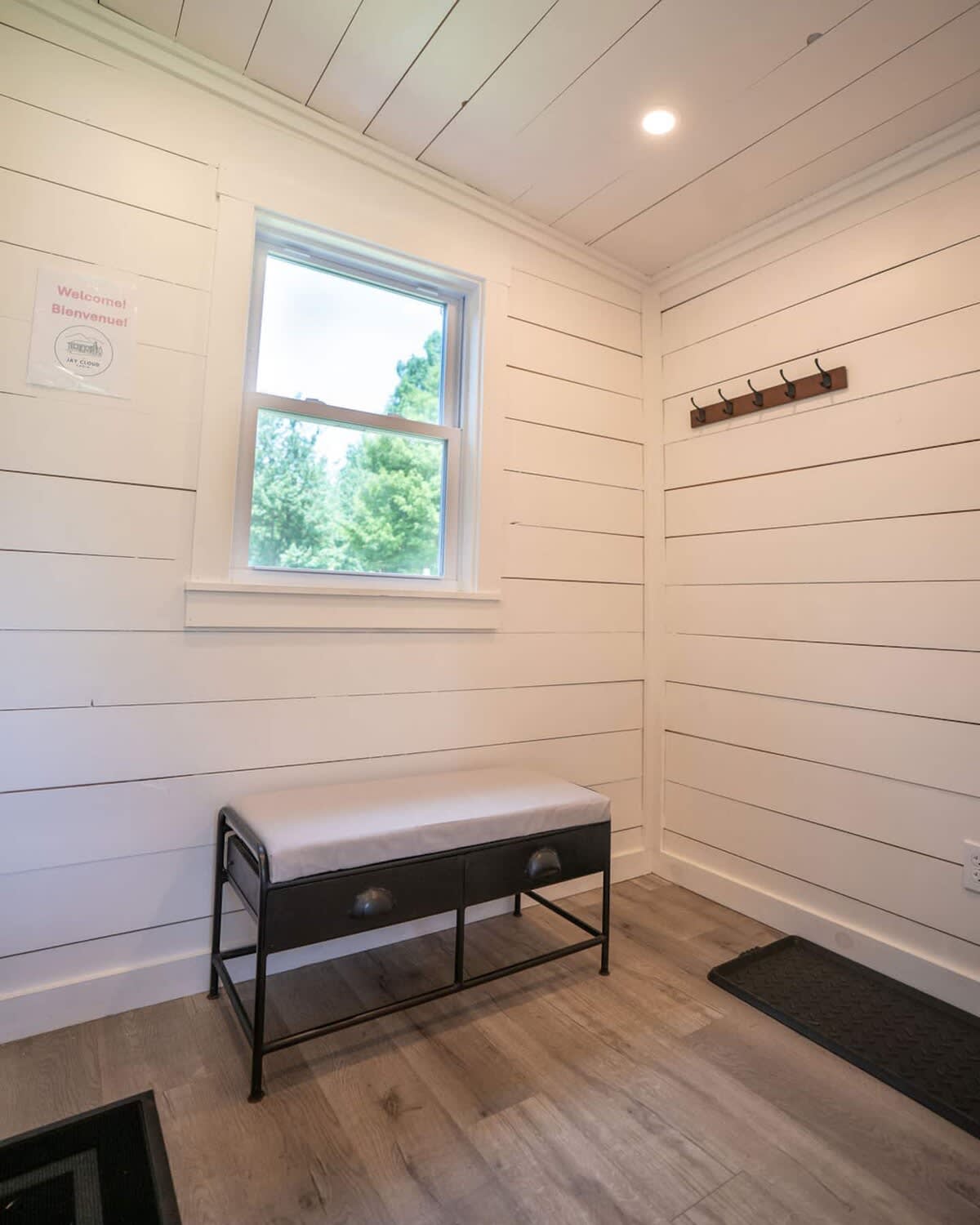 A bright and inviting mudroom features white shiplap walls and ceiling, with a window offering a view of lush trees. A black metal bench with a cushioned seat provides a place to sit, and a coat rack with hooks is mounted on the wall.