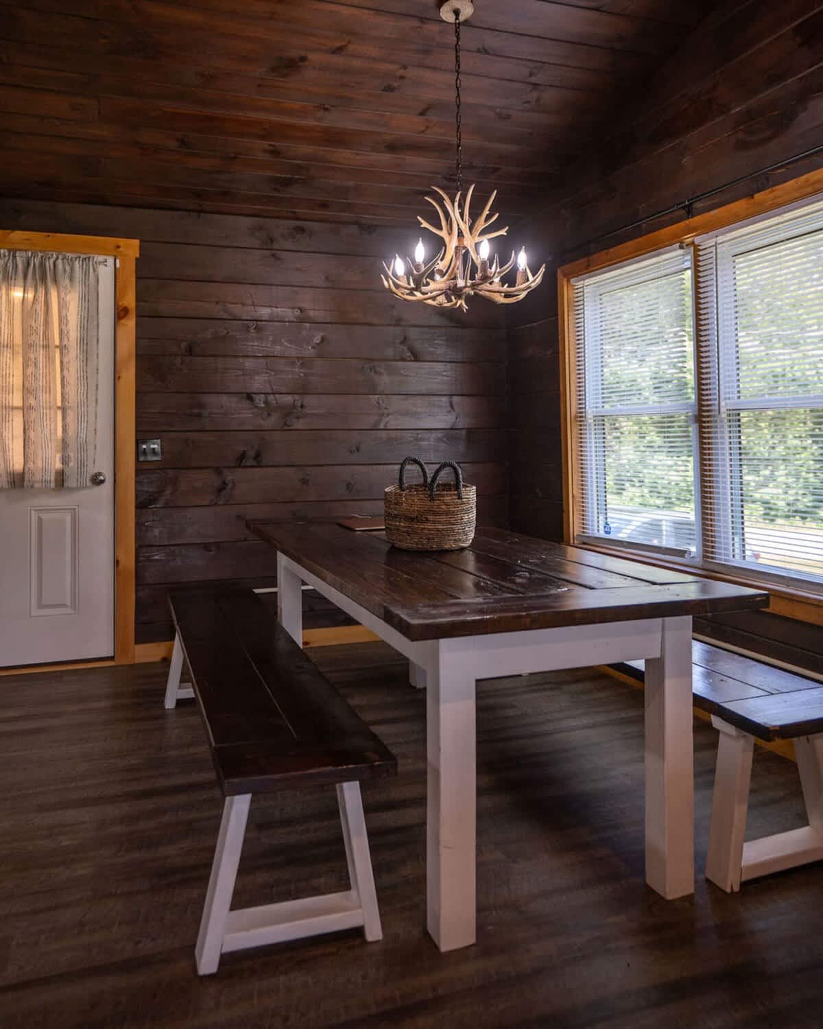 Rustic dining area with dark wood paneled walls and ceiling, featuring a long wooden dining table with white legs and two matching benches, illuminated by an antler chandelier.