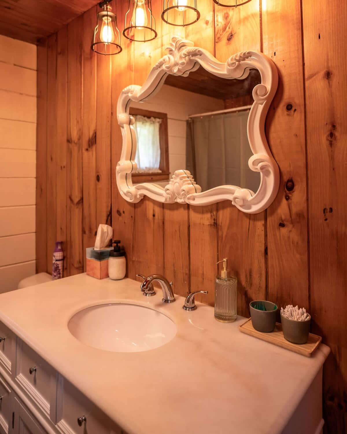 A close-up of the bathroom vanity showcases a white sink with chrome fixtures set in a light countertop, surrounded by various toiletries. An ornate white framed mirror reflects a shower curtain, illuminated by industrial-style light fixtures above.