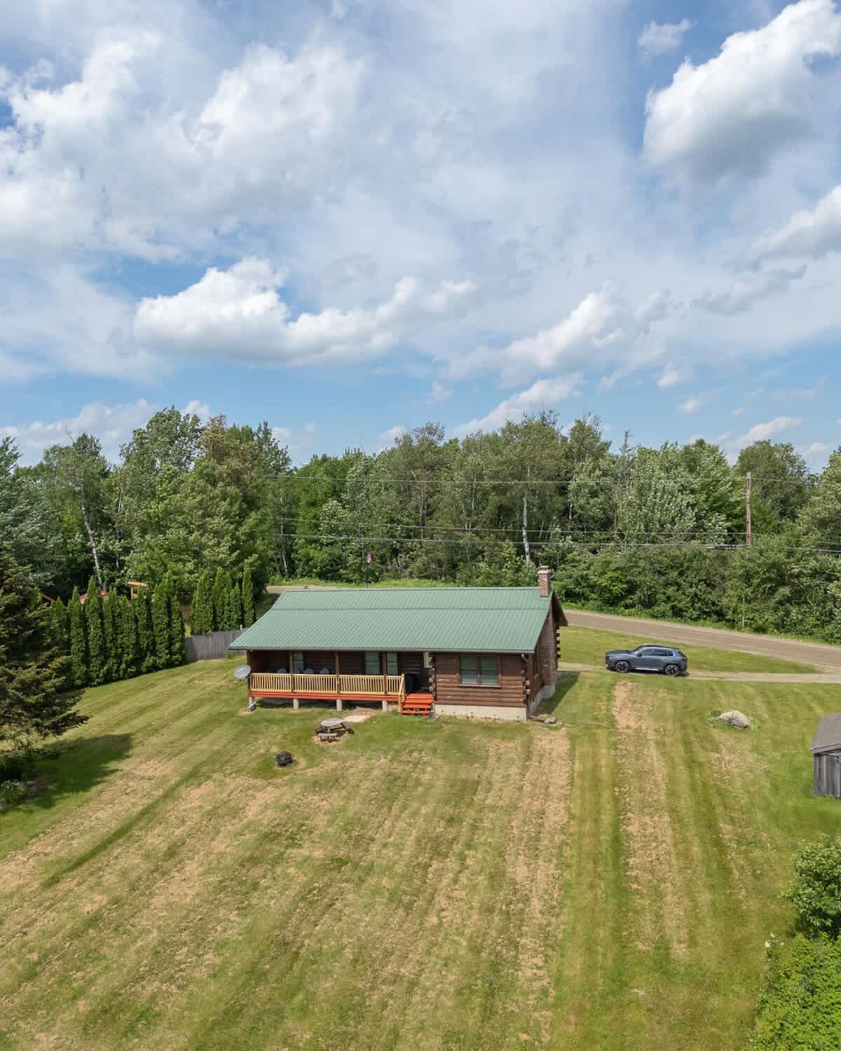 Aerial view of a log cabin with a green metal roof and a covered porch, situated on a large grassy property with trees and a car parked nearby, under a partly cloudy sky.