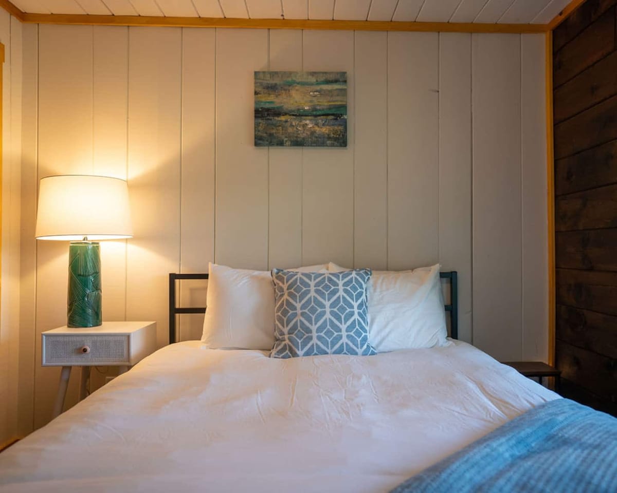 Close-up of a queen-sized bed with white bedding and a blue patterned pillow, flanked by a white nightstand with a tall green lamp. The room has white vertical plank walls.