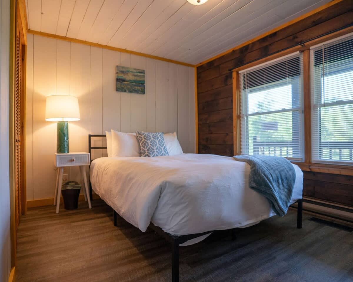 Queen-sized bed with white bedding and a blue throw in a bedroom with white vertical plank walls and a dark wood accent wall. Two windows with blinds offer natural light.