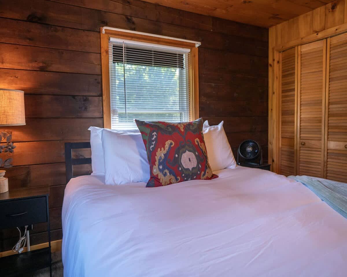 Close-up of a queen-sized bed with white bedding and a vibrant patterned pillow in a bedroom with dark wood plank walls. A dark nightstand with a fan is beside the bed.
