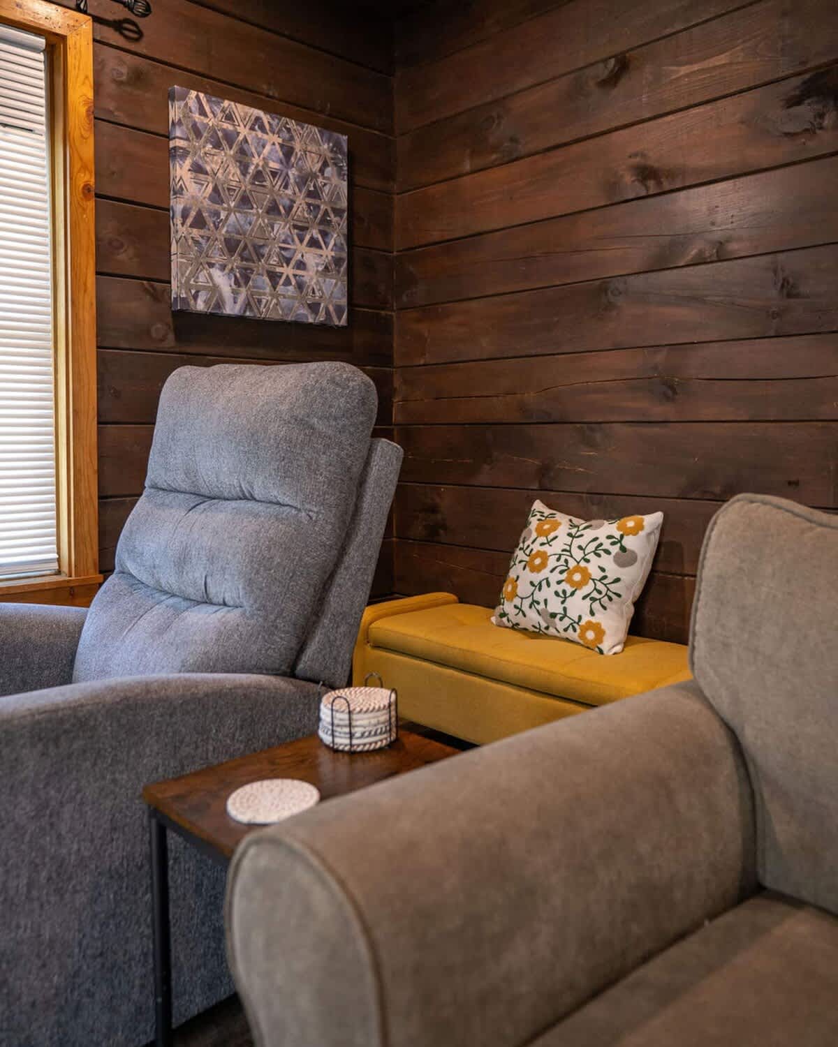 A comfortable living room seating area features dark wood paneled walls, with a grey recliner and a beige sofa separated by a small wooden side table. A vibrant yellow storage bench with a decorative pillow adds a pop of color and extra seating.
