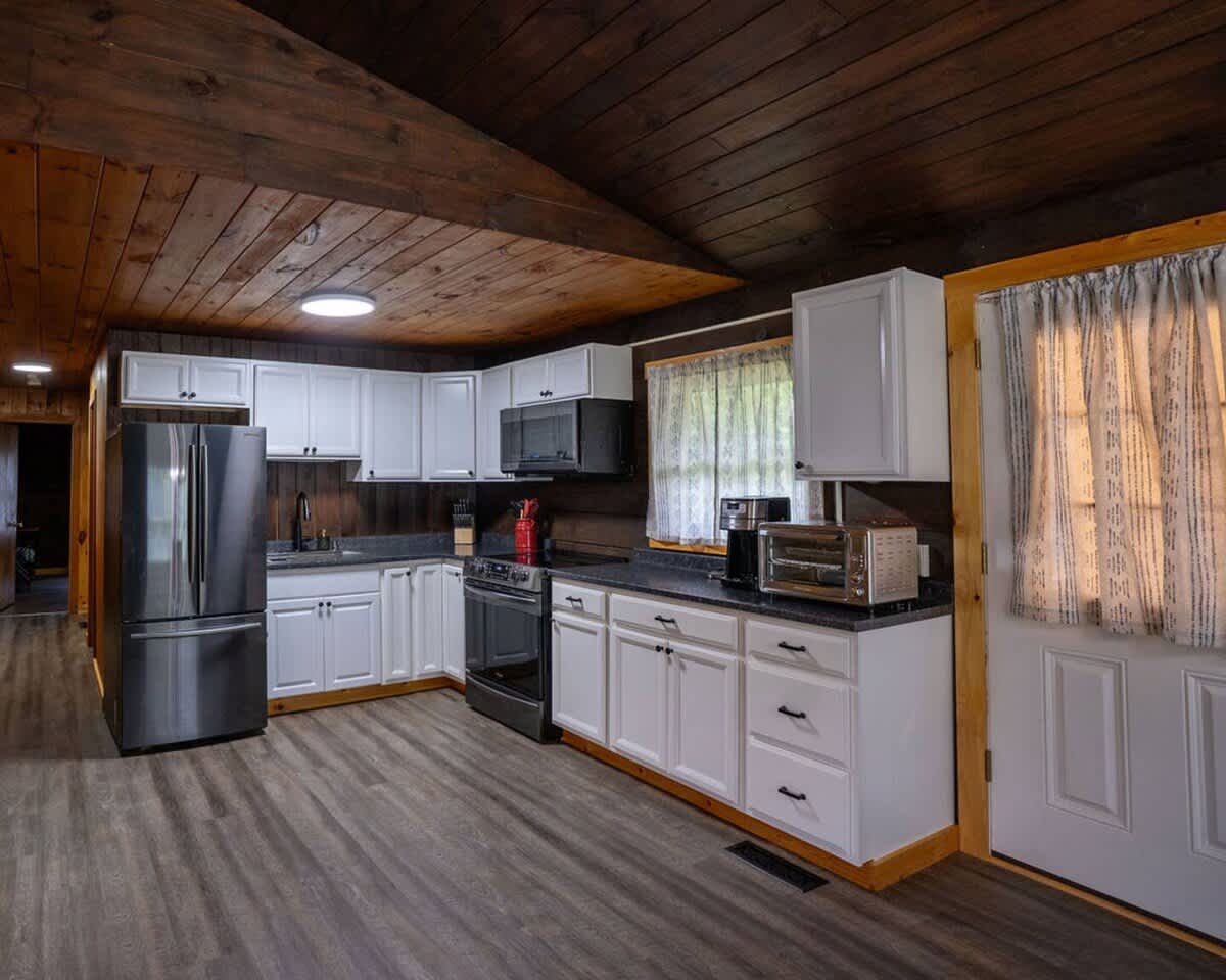 A full view of the kitchen reveals dark wood paneled walls and ceiling, contrasting with white shaker-style cabinets and black countertops. Stainless steel appliances, including a refrigerator, stove, and microwave, are integrated into the space, alongside a window and an exterior door.