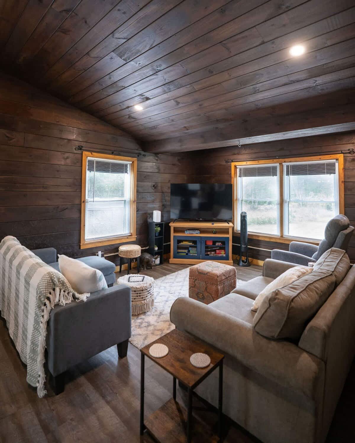 A cozy living room with dark wood paneled walls and ceiling is furnished with a grey sectional sofa and a matching armchair, centered around a patterned area rug. A large flat-screen TV sits on a blue media console, flanked by two windows with blinds.
