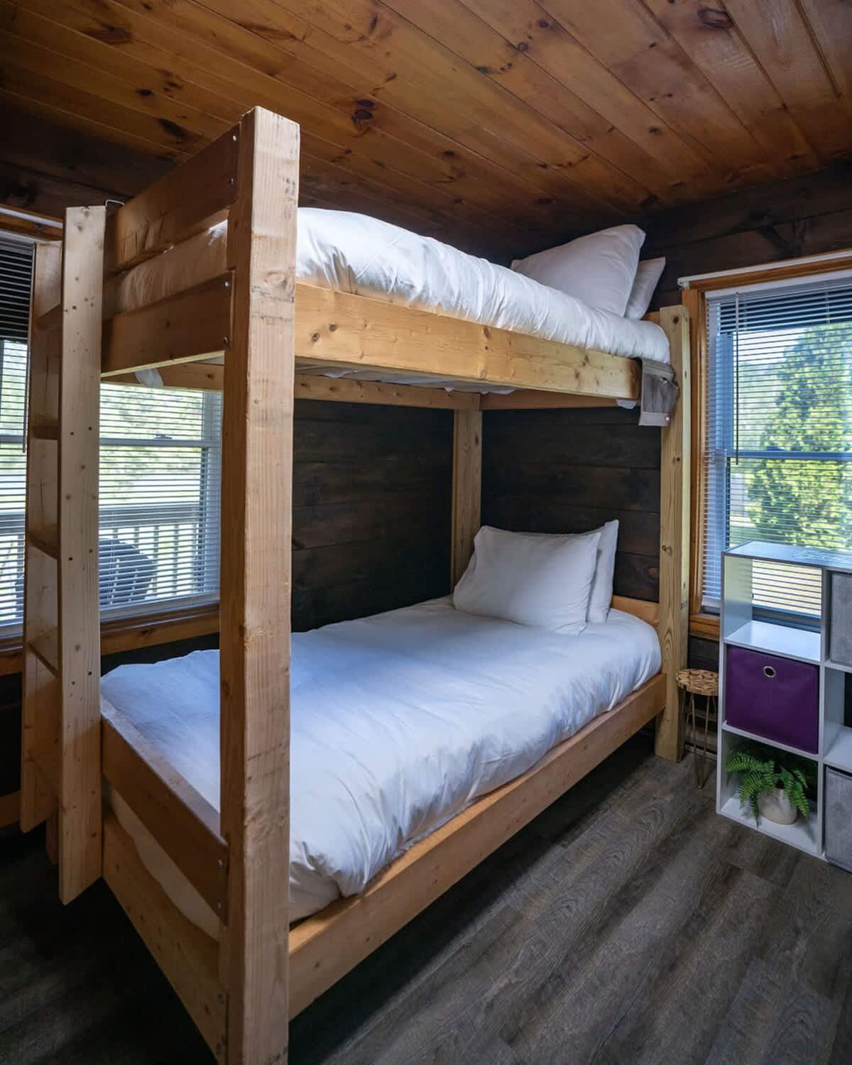 A cozy bedroom features a sturdy wooden bunk bed with crisp white bedding, set against dark wood paneled walls and ceiling. Two windows with blinds provide natural light, complemented by a small white shelving unit with a purple storage bin.