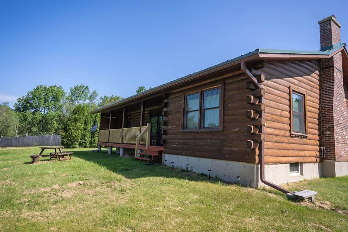 Side view of a rustic log cabin with a covered porch, a brick chimney, and a green metal roof, situated on a grassy lawn under a clear blue sky.