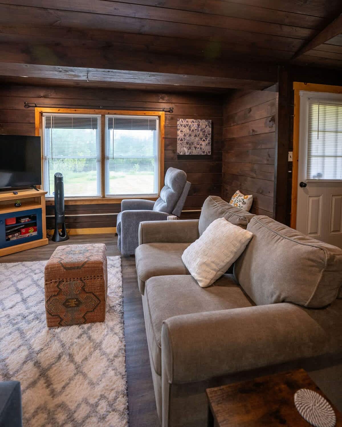 Living room with dark wood paneled walls and ceiling, a gray sofa, a patterned ottoman, and large windows overlooking a green outdoor area.