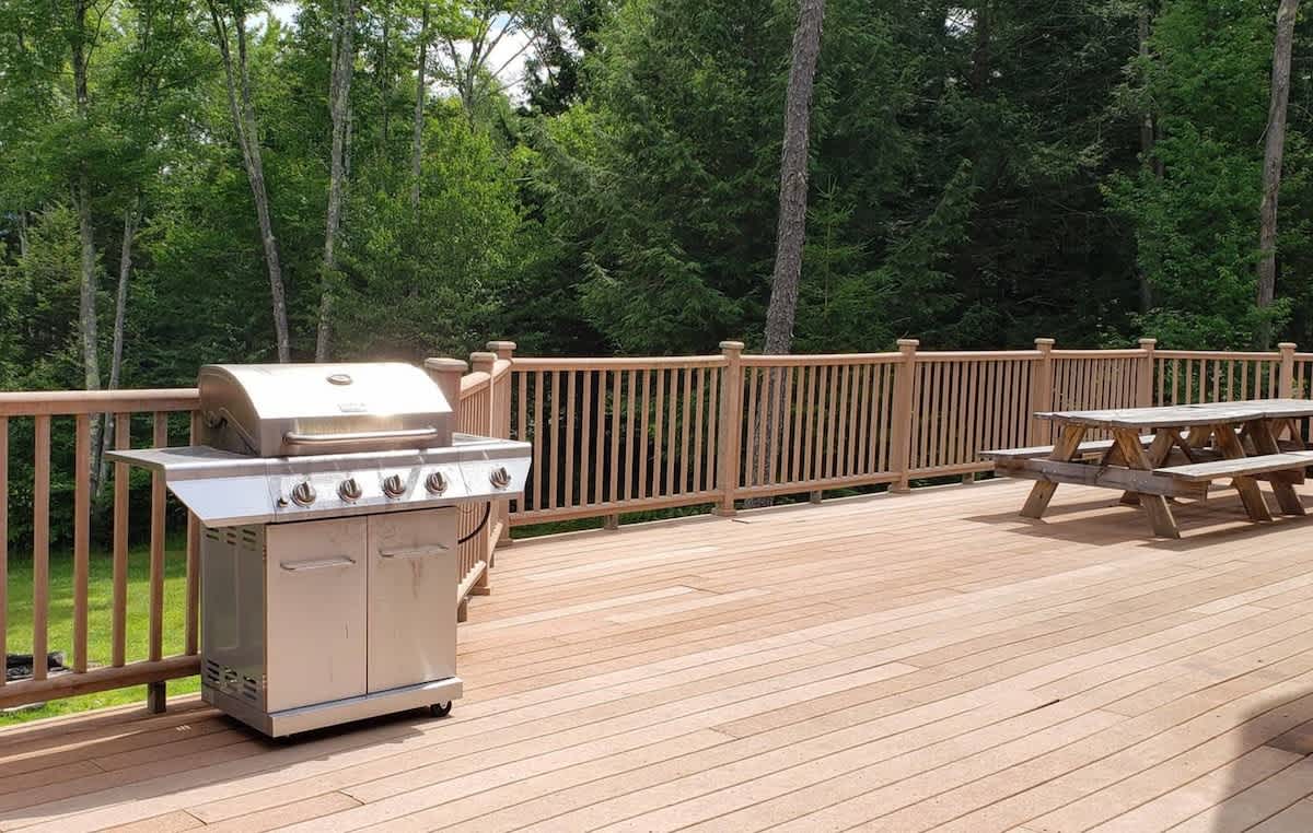 A spacious wooden deck with a sturdy railing, featuring a large stainless steel BBQ grill and a wooden picnic table. The deck overlooks a lush green forest.
