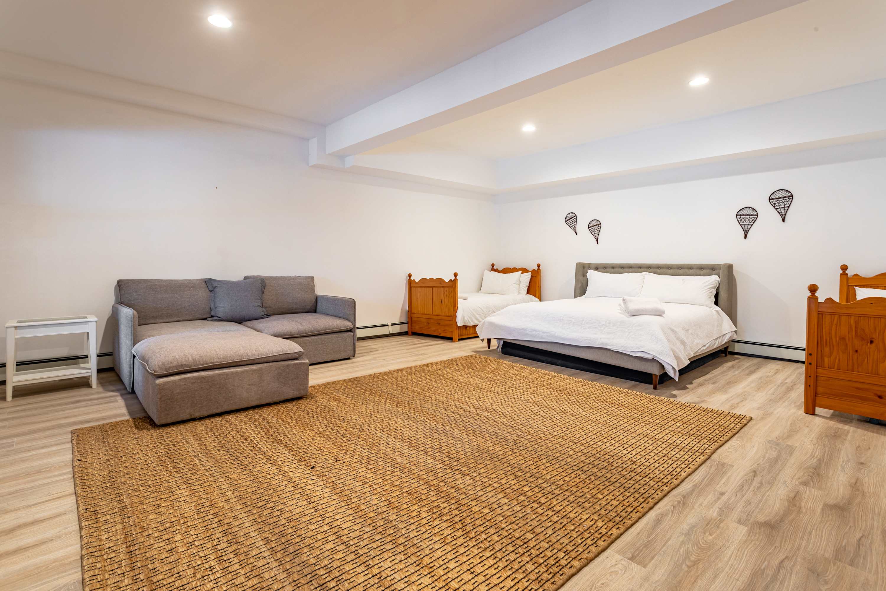 A large bedroom with light wood-look flooring and a woven rug, featuring a queen-sized bed, two twin beds, and a grey sectional sofa with an ottoman. The room is brightly lit with recessed lighting.