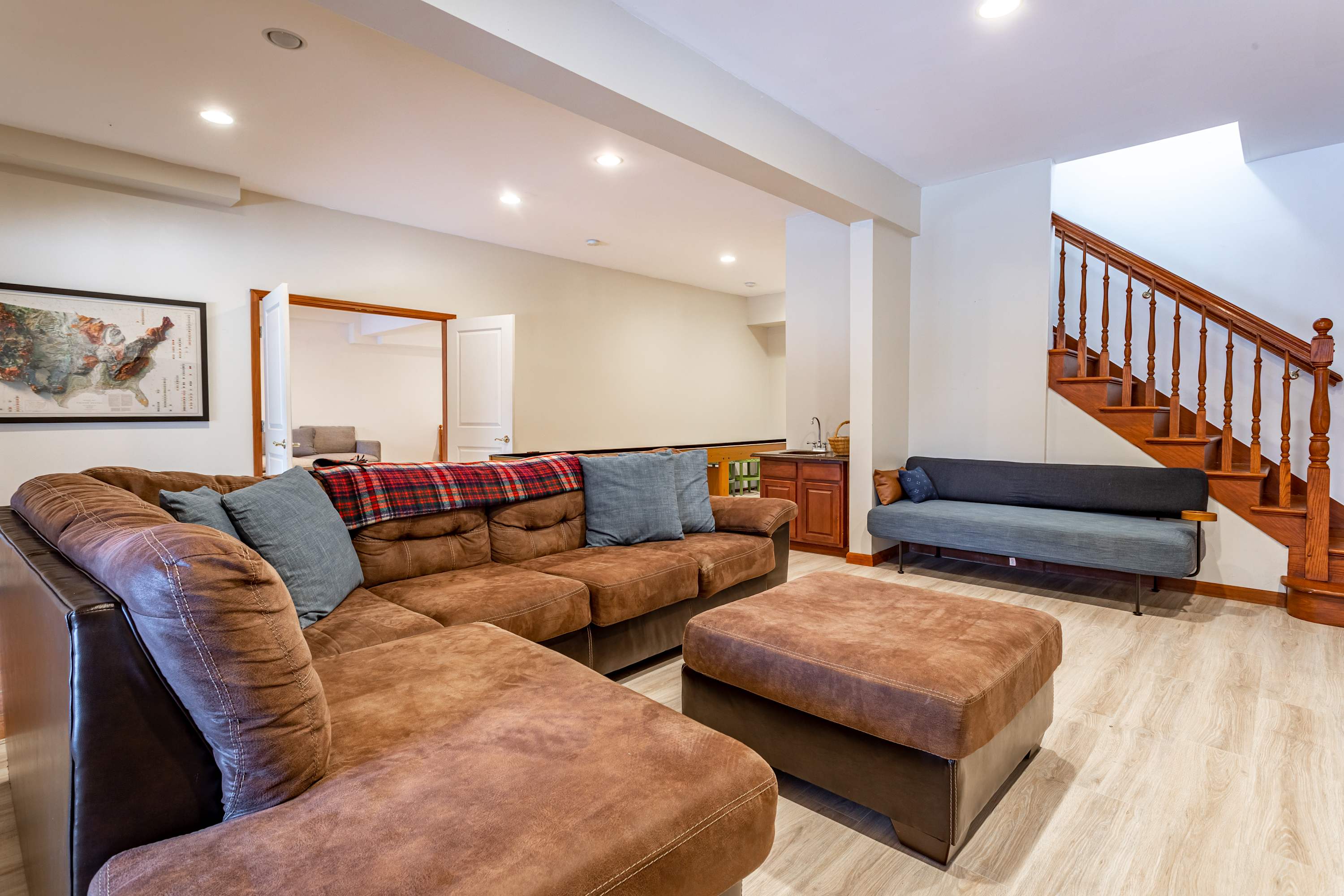 A spacious living area with light wood-look flooring, featuring a large brown sectional sofa with blue pillows, a grey daybed, and a wooden staircase leading upstairs. A small wet bar is visible in the background.