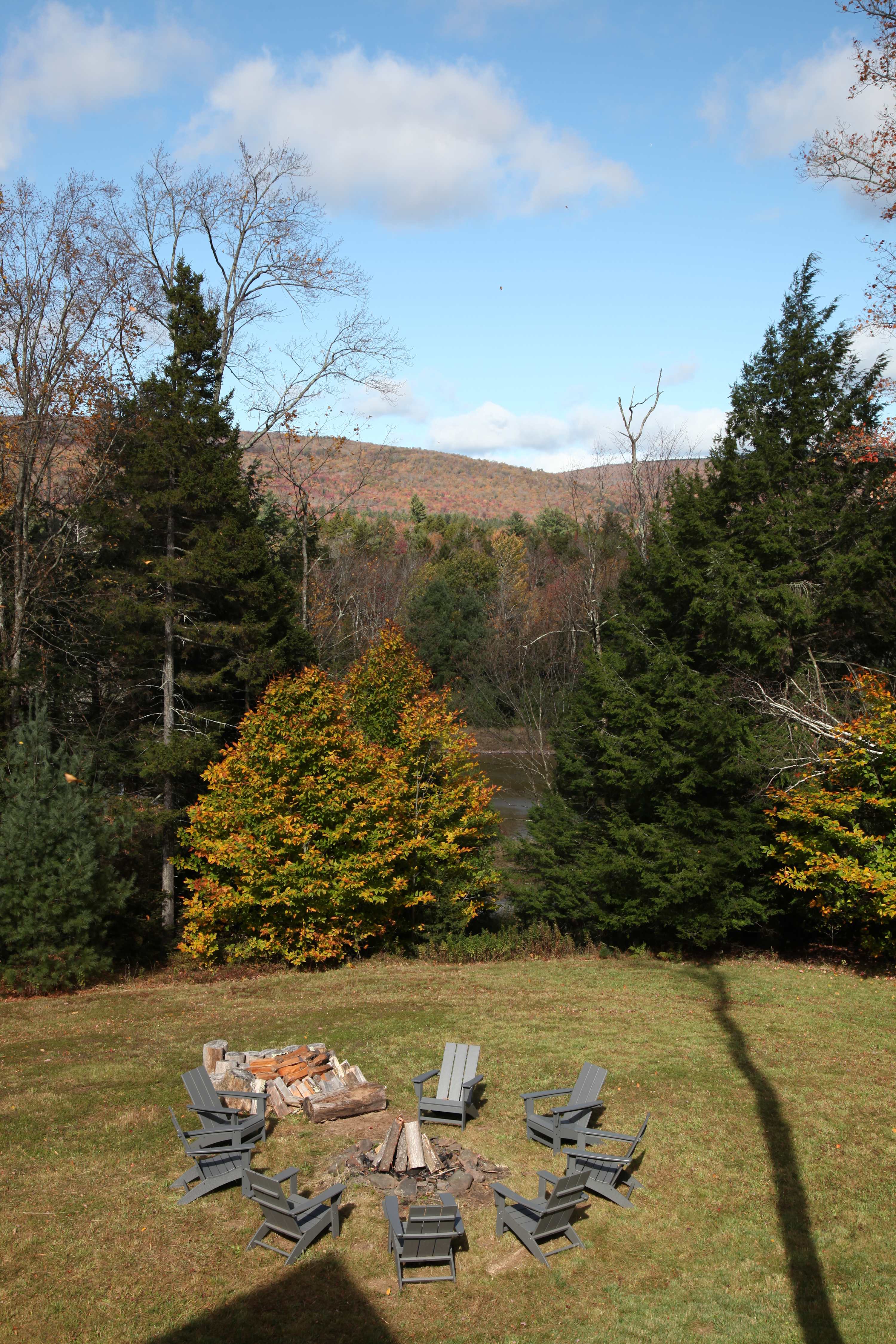 An inviting fire pit surrounded by grey Adirondack chairs sits on a lush green lawn, overlooking a serene body of water and rolling hills covered in a mix of evergreen and colorful autumn trees. A long shadow stretches across the grass.