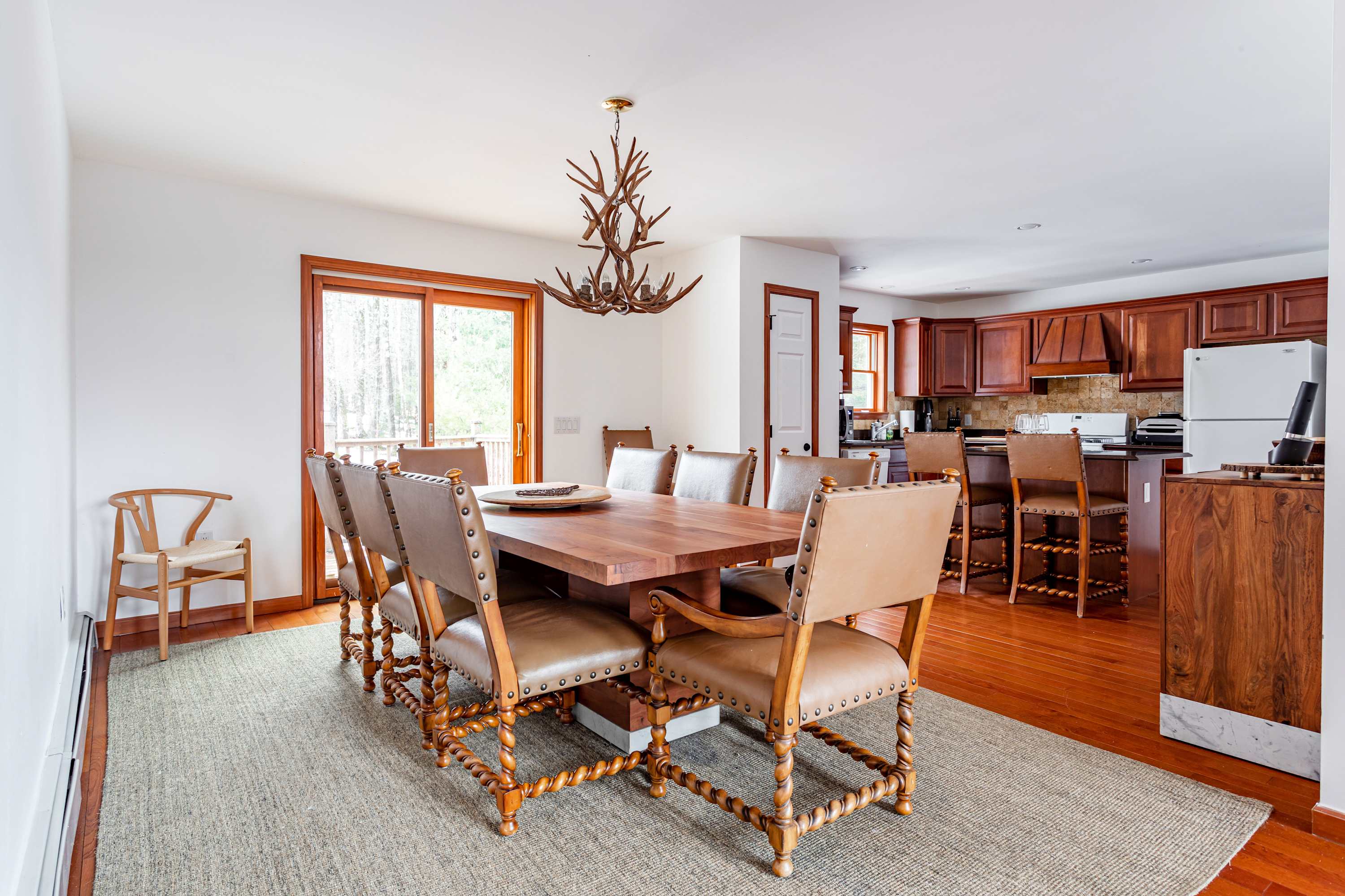 A formal dining room features a large wooden table with eight brown leather chairs, illuminated by an antler-style chandelier. Polished hardwood floors and a light area rug complement the white walls, with a sliding glass door leading to the outdoors.