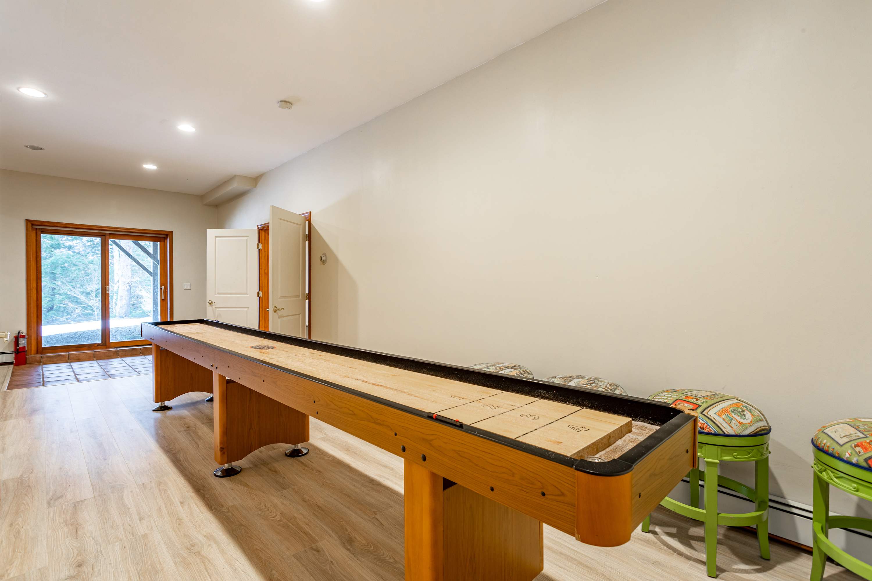 A bright game room with light wood-look flooring, featuring a long wooden shuffleboard table and two green stools with patterned cushions. A glass door leads to the outside, revealing trees.