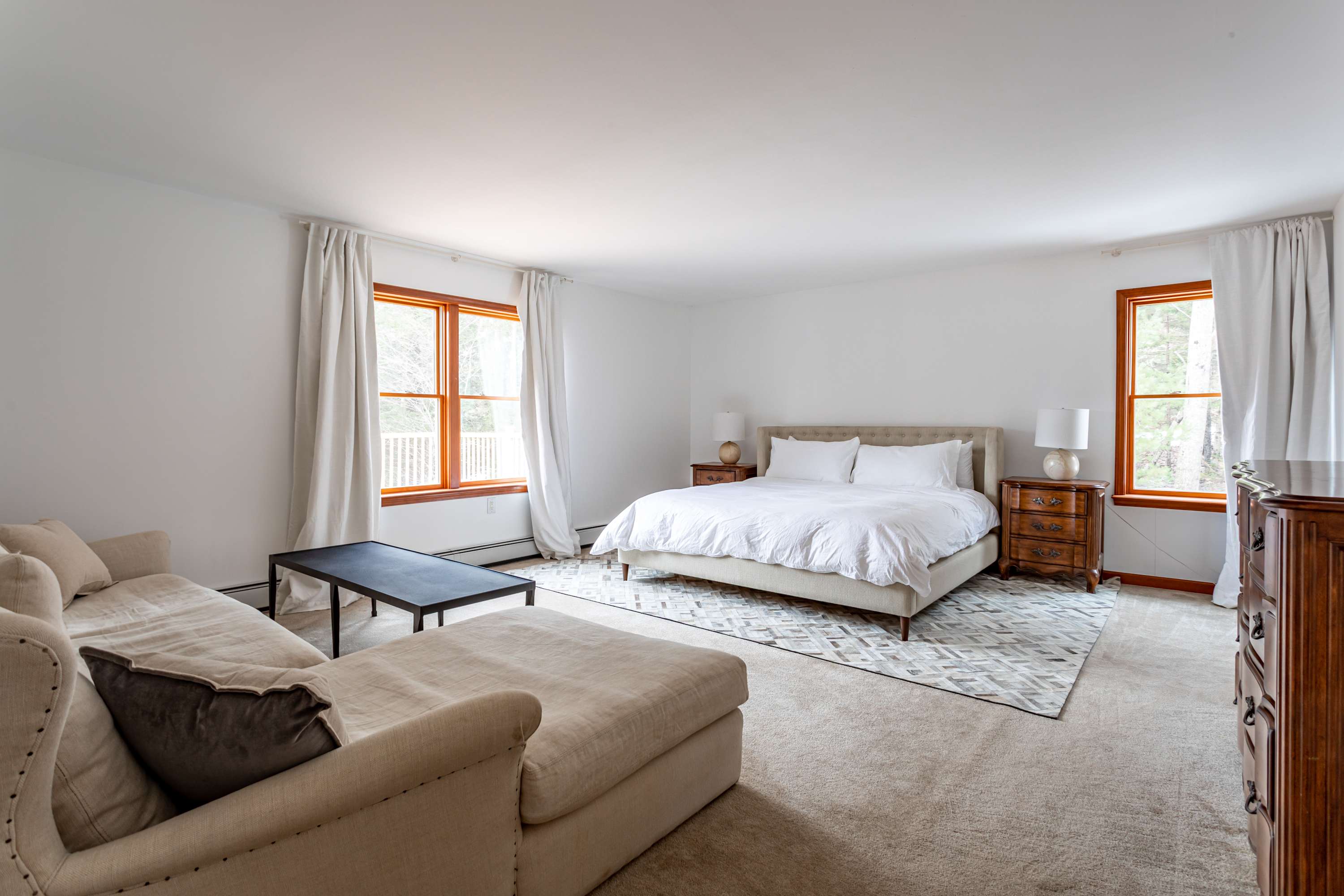 A spacious master bedroom featuring a king-sized bed with a tufted headboard, crisp white bedding, and two wooden nightstands with lamps, complemented by a light-colored sectional sofa and a black coffee table on a patterned area rug.