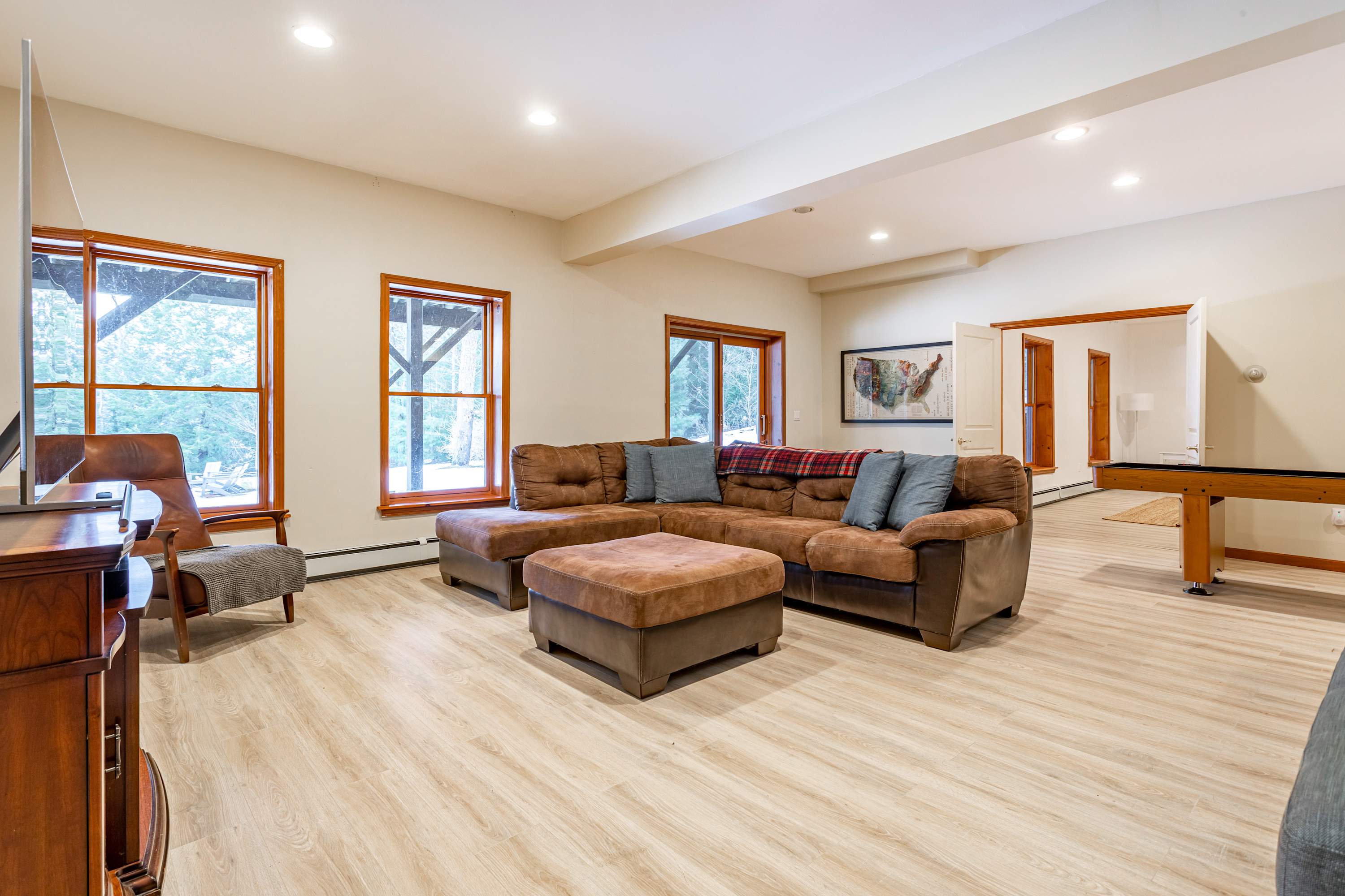 Expansive living room with light wood-look flooring and large windows offering views of lush trees, furnished with a large brown sectional sofa, ottoman, and a TV console. The room has recessed lighting and light-colored walls.