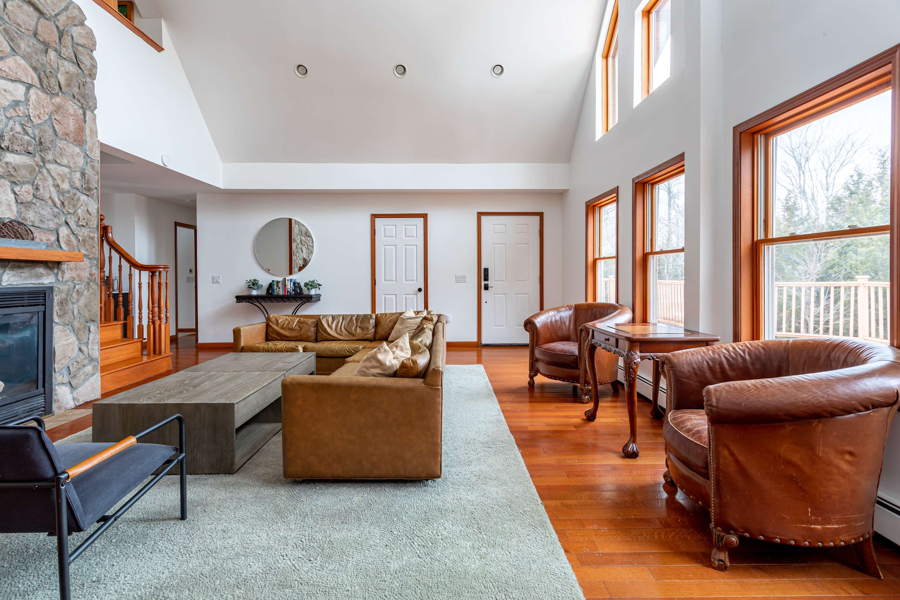 A bright living room with vaulted white ceilings and large windows showcasing outdoor views. It features a brown leather sectional sofa, a modern wooden coffee table, and two brown leather armchairs on a light area rug, all set on polished hardwood floors.