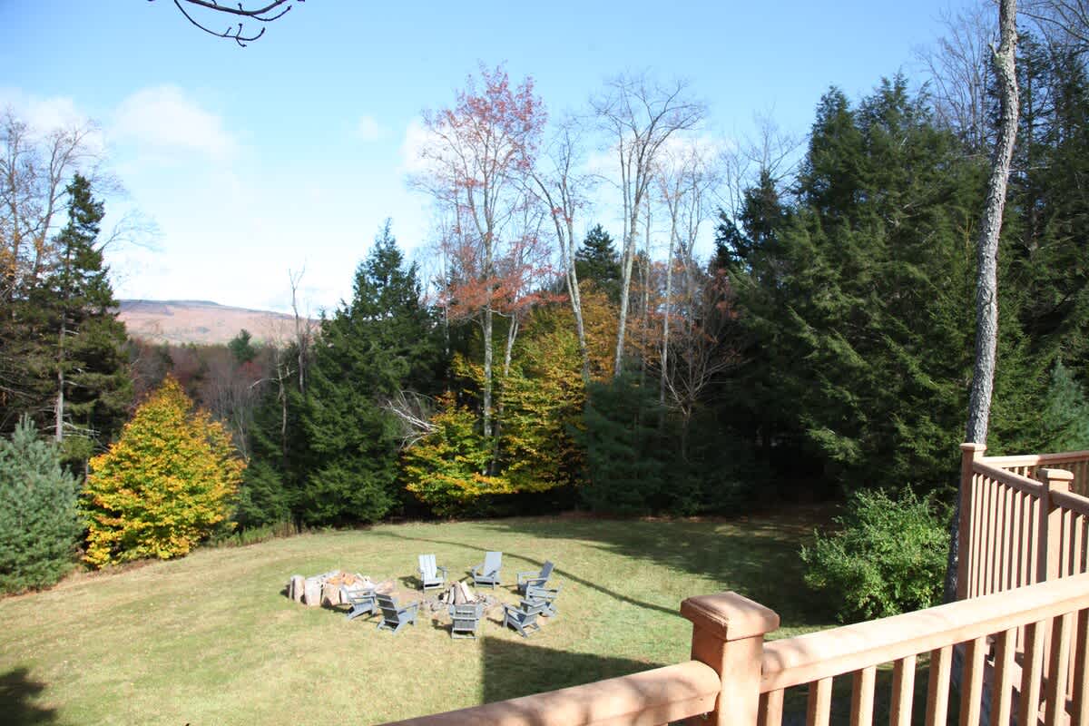 A spacious grassy yard features a circular fire pit surrounded by numerous grey Adirondack chairs, set against a backdrop of vibrant autumn trees and distant mountains under a partly cloudy sky. A wooden deck railing is visible in the foreground.