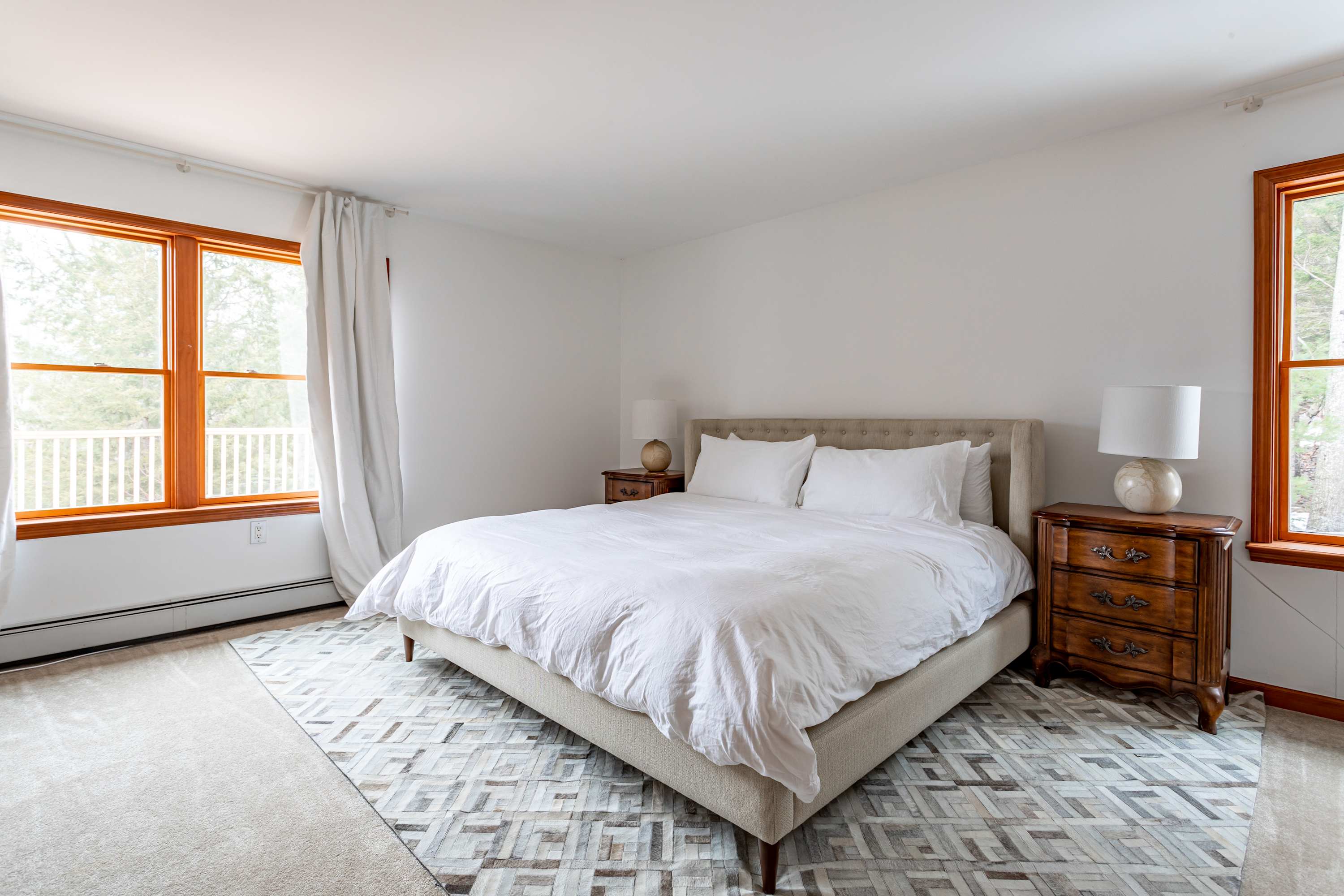 A close-up view of the king-sized bed in the master bedroom, showcasing its light-colored tufted headboard, pristine white bedding, and the two wooden nightstands with matching lamps, all centered on a geometric patterned rug.
