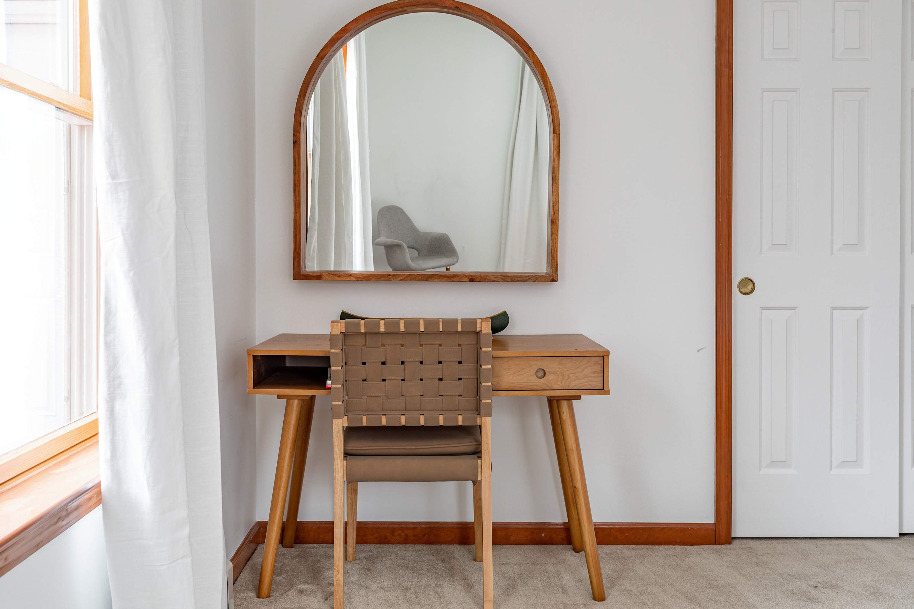 A stylish vanity or desk area featuring a light wood desk with a woven back chair, topped with an arched wooden mirror, positioned next to a window with white curtains and a white closet door.