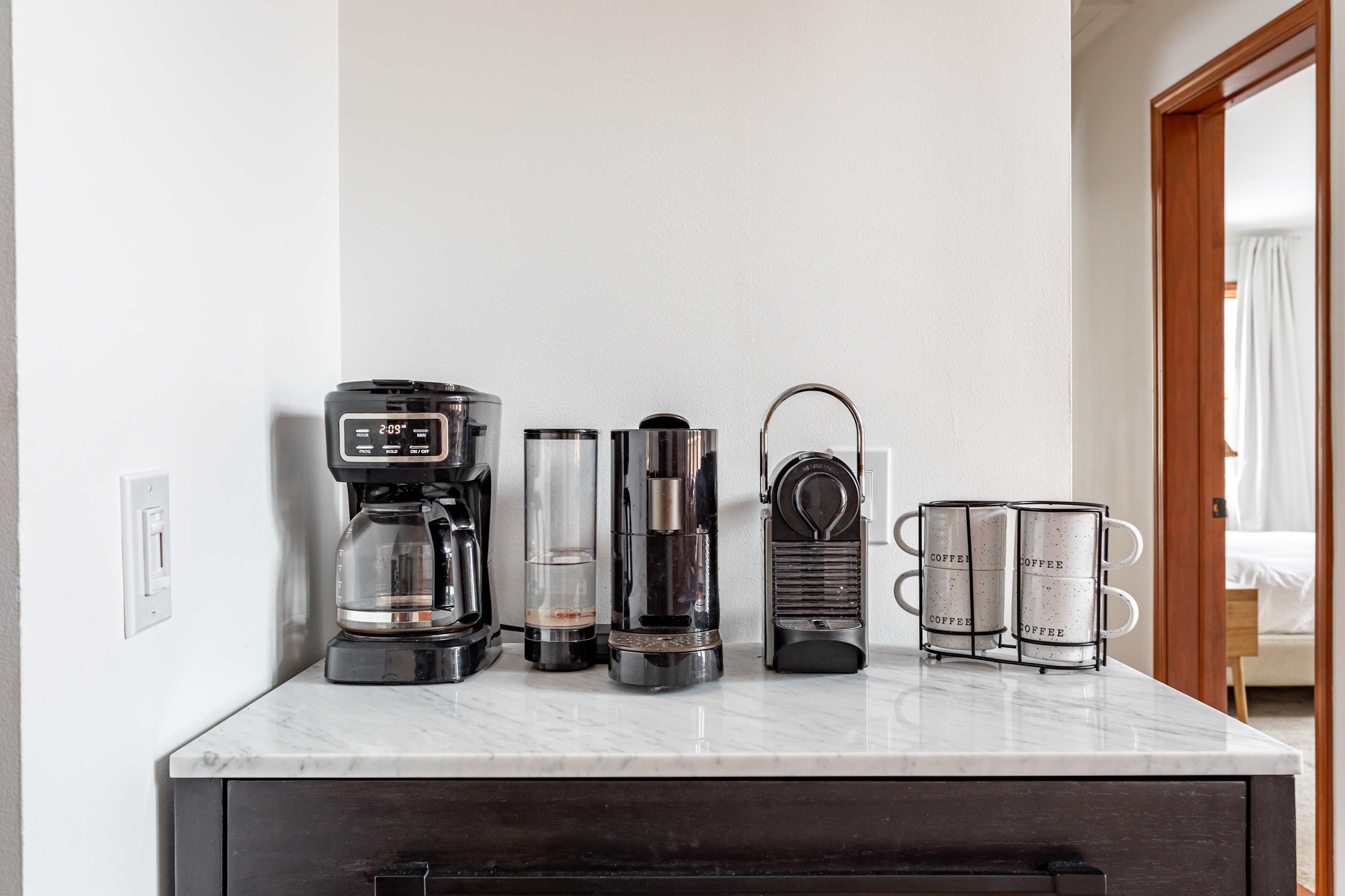 A dedicated coffee station features a white marble countertop with various coffee makers, including a drip coffee machine and two Nespresso machines. A stack of white coffee mugs completes this organized beverage area.