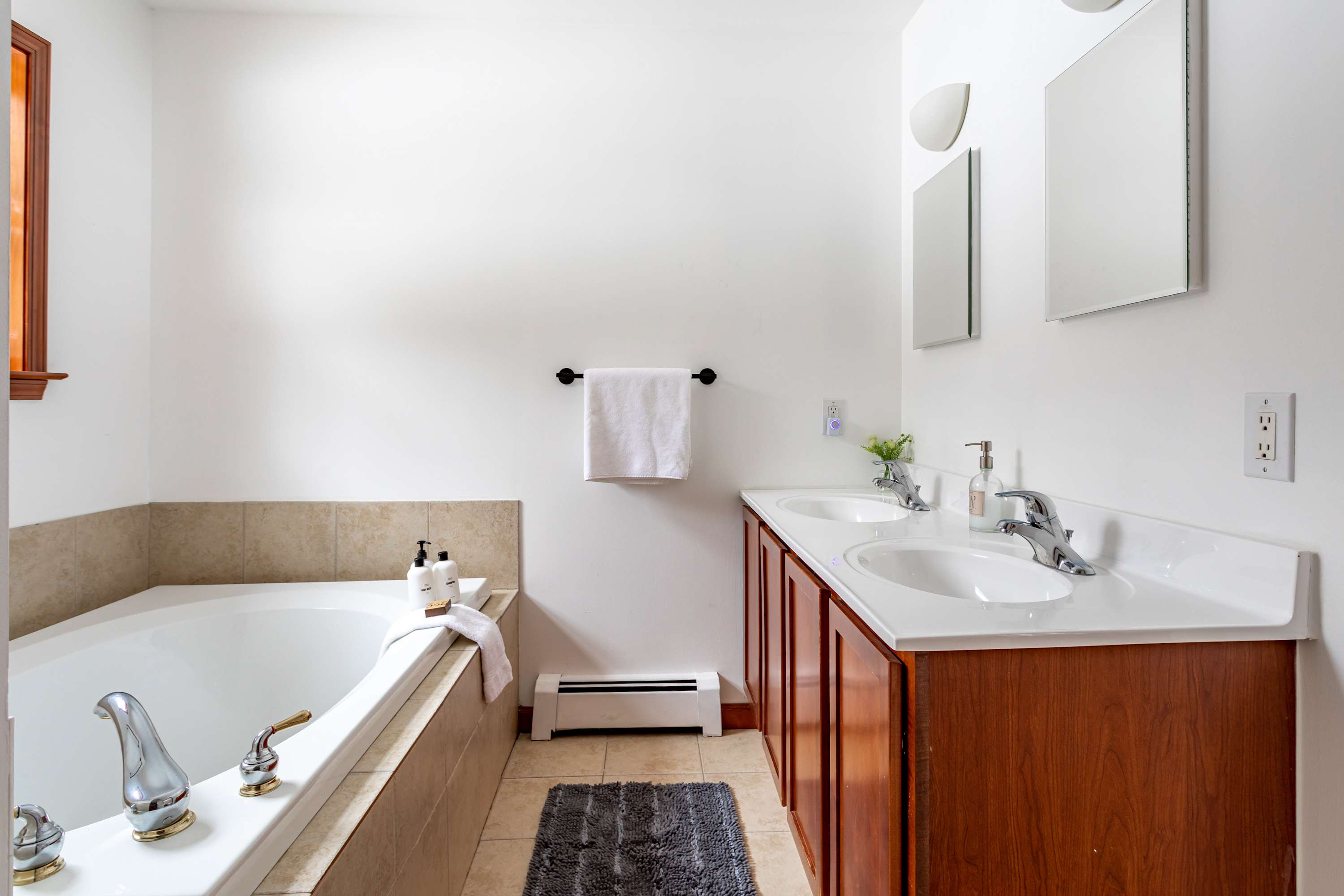 A bright master bathroom featuring a large corner soaking tub with beige tile surround, a double vanity with wooden cabinets and white countertops, and two mirrors, set against white walls and a tiled floor.