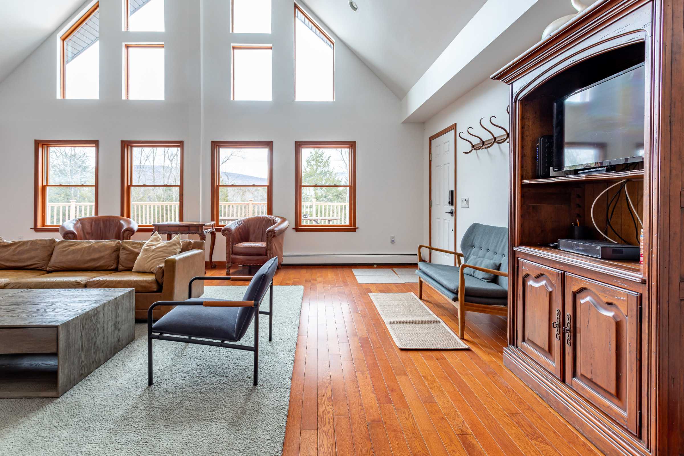A spacious living area with high vaulted ceilings and multiple large windows offering views of the outdoors. The room features polished hardwood floors, a light area rug, a brown leather sofa, and a dark wooden entertainment cabinet.