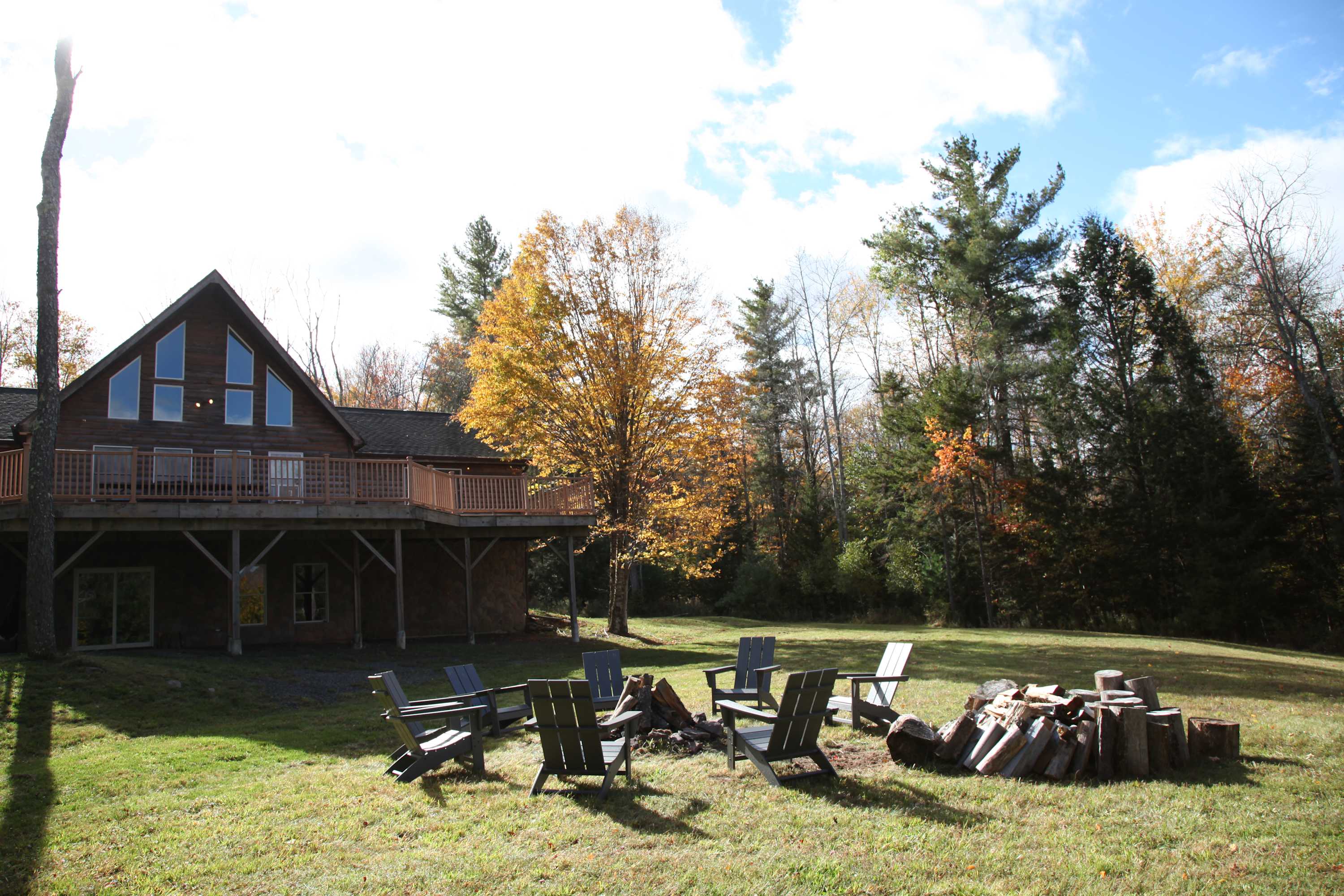 Outdoor fire pit area with several Adirondack chairs arranged around a central fire pit and a stack of firewood. The wooden cabin with its large deck is visible in the background, surrounded by autumn trees.