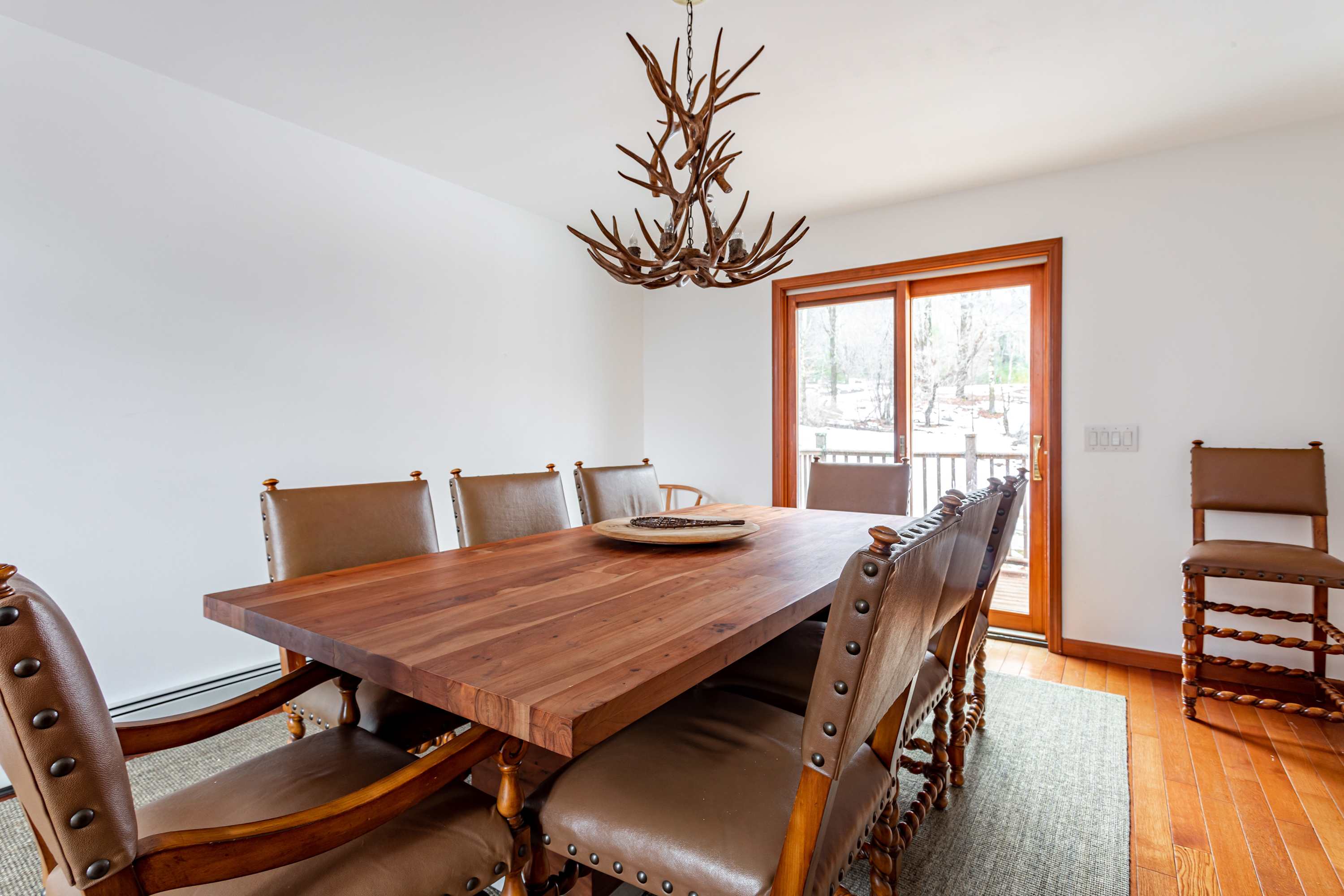 Elegant dining room featuring a large rustic wooden table with seating for eight, complemented by leather-upholstered chairs with nailhead trim. A unique antler-style chandelier hangs above, and a sliding glass door leads to an outdoor area.