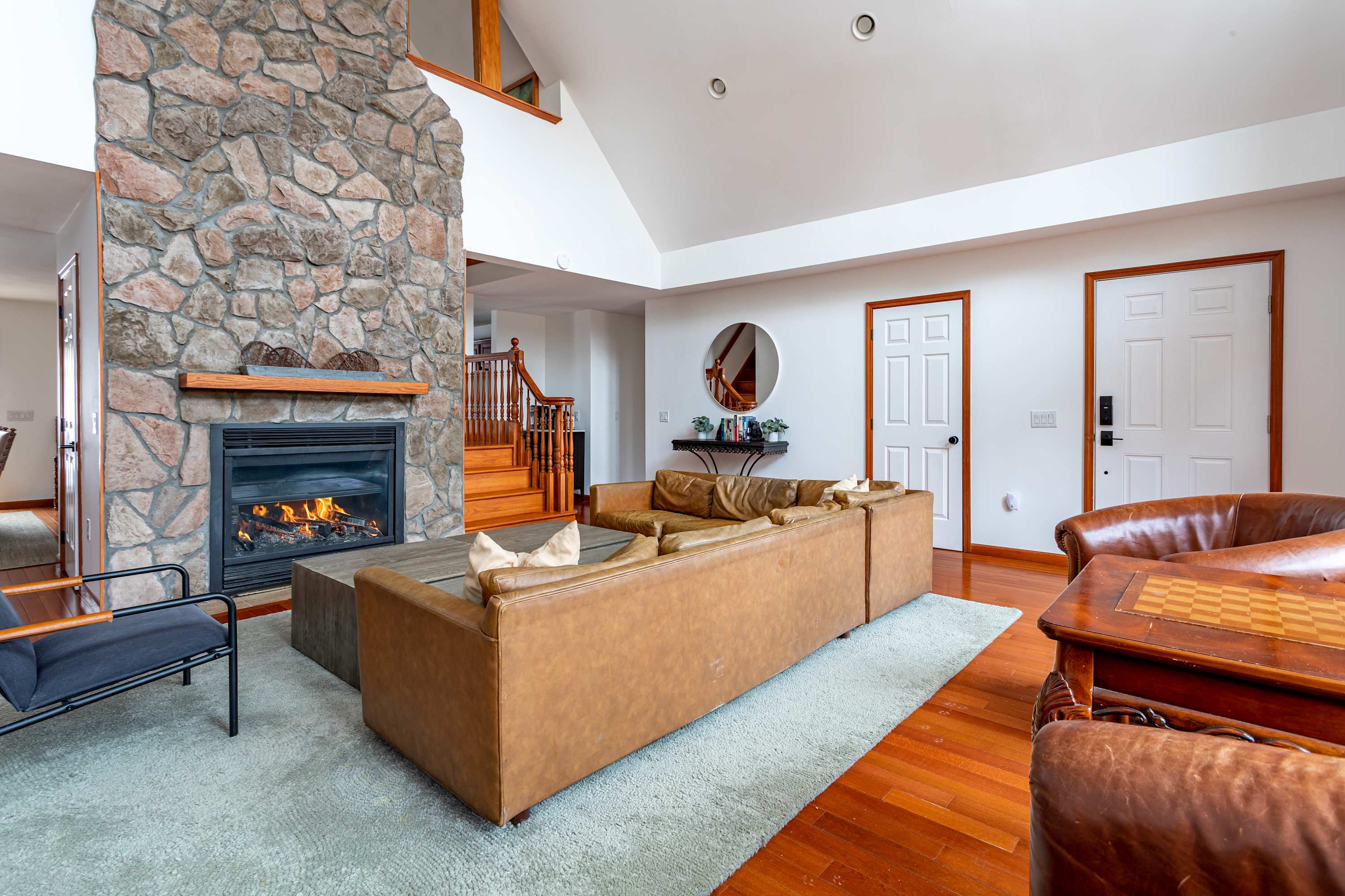 A cozy living room features a large L-shaped brown leather sofa on a light rug, facing a grand stone fireplace with a lit fire. Hardwood floors and white walls with high ceilings create an open, inviting atmosphere.