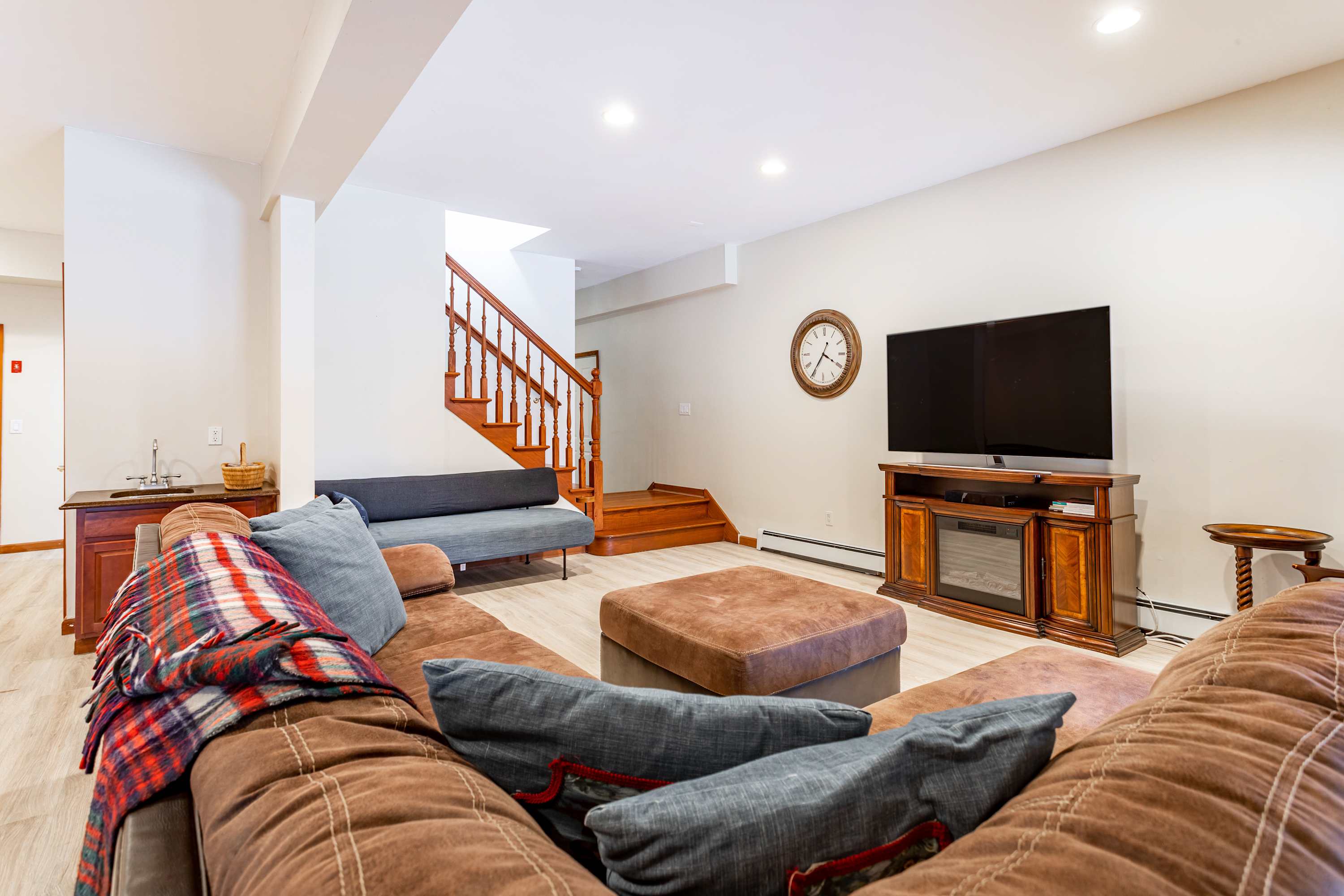 Basement media room with a large brown sectional sofa, a flat-screen TV on a wooden console, and light-colored flooring. A wooden staircase leads up to the main level, and a small wet bar is visible in the corner.