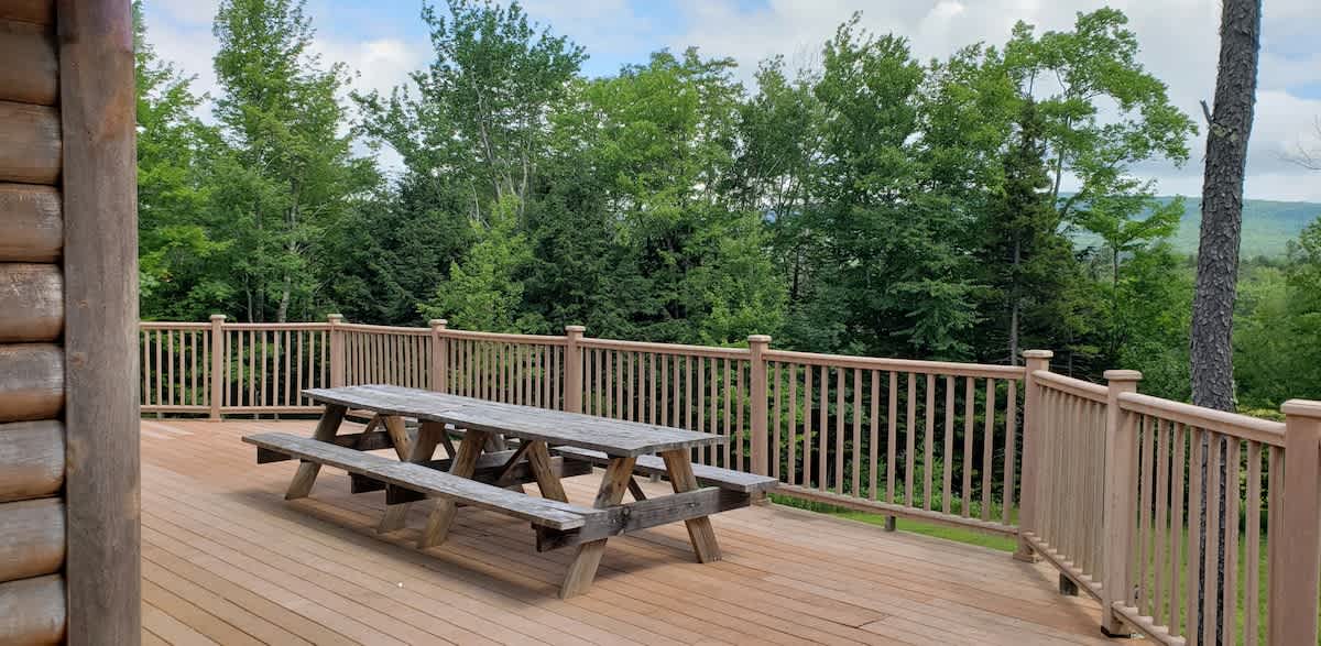 A spacious outdoor deck features a rustic wooden picnic table with benches, surrounded by a light-colored wooden railing. Lush green trees and distant mountains under a partly cloudy sky provide a scenic backdrop.