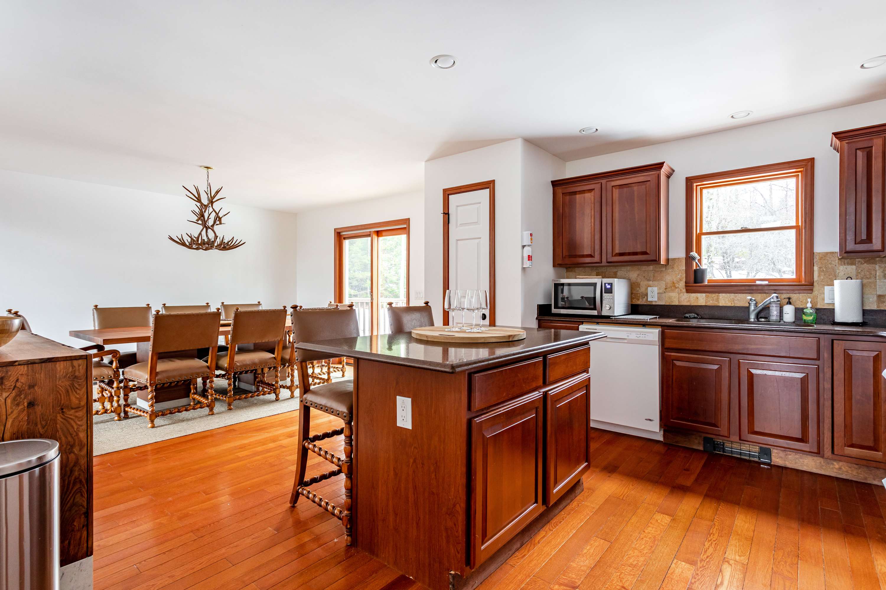 A view of the kitchen and dining area, showing the wooden kitchen island with bar stools in the foreground and the large dining table with leather chairs in the background. The space features warm hardwood floors and dark wooden cabinetry.