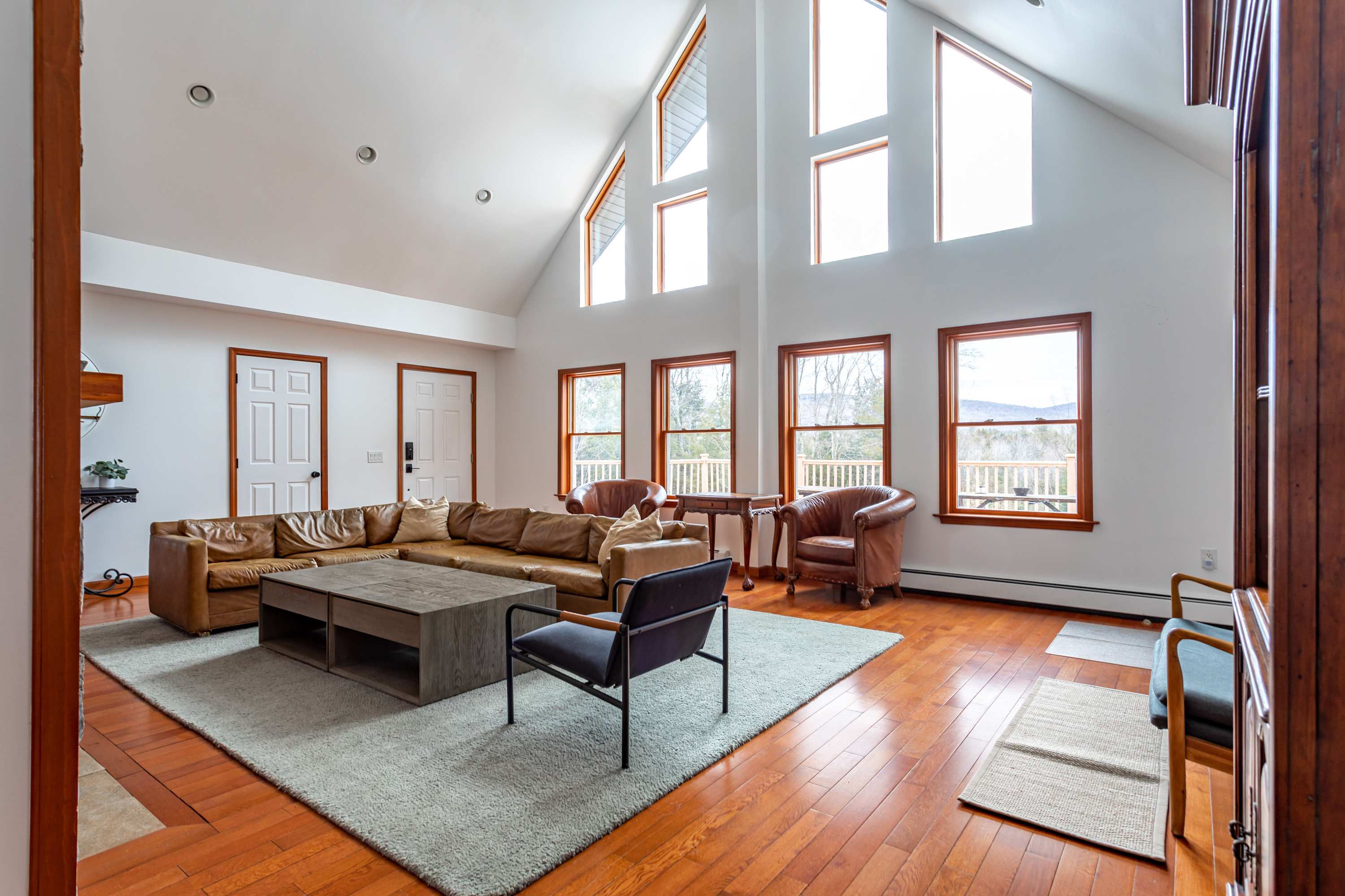 Spacious living room with light brown leather sectional sofa, a large grey coffee table, and warm hardwood floors. Tall vaulted ceilings and numerous windows offer abundant natural light and views of the surrounding landscape.