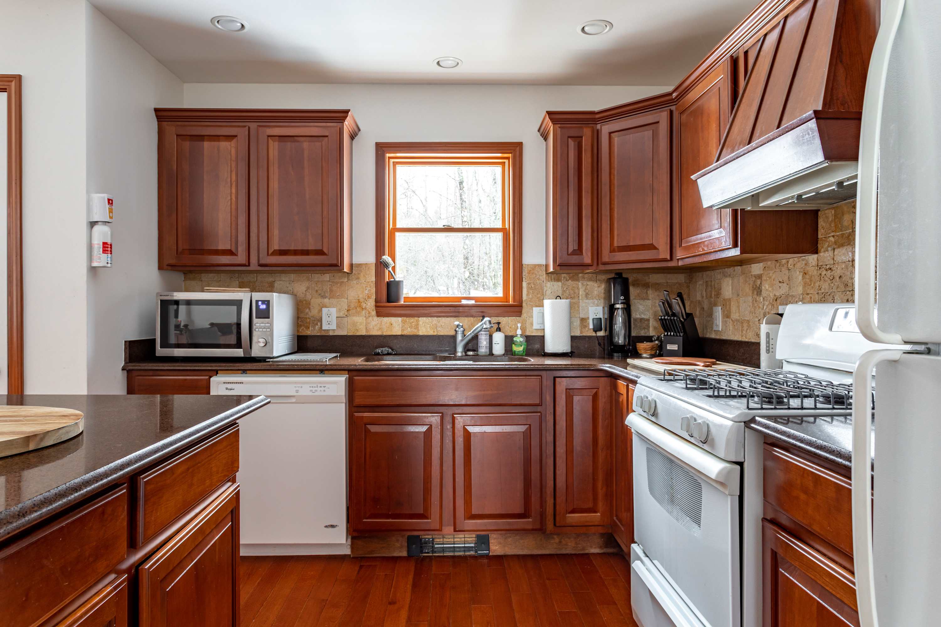 A detailed view of the kitchen counter area with dark countertops, wooden cabinets, and a tiled backsplash. A white microwave, dishwasher, and gas range are visible, along with a window overlooking green trees.