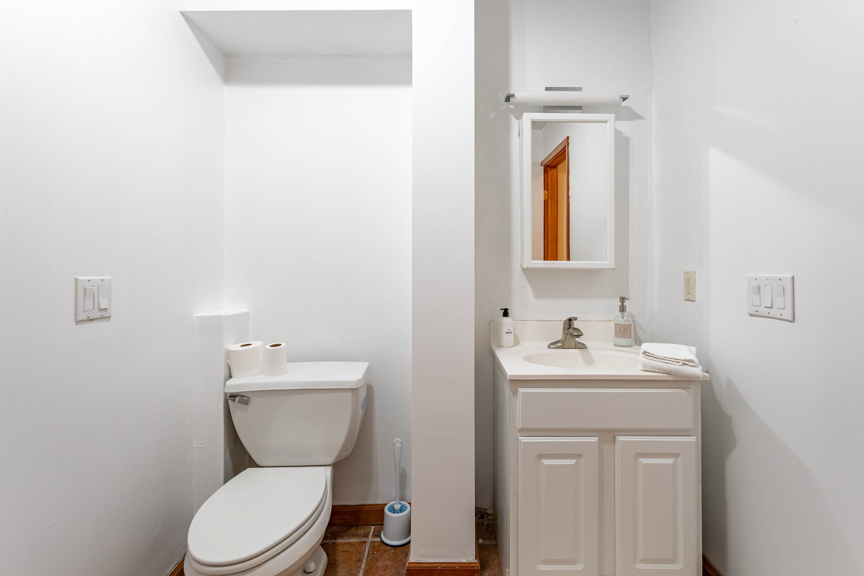 A compact white bathroom featuring a toilet with toilet paper rolls on top and a white vanity with a sink, mirror, and basic toiletries. The floor is tiled in a reddish-brown.