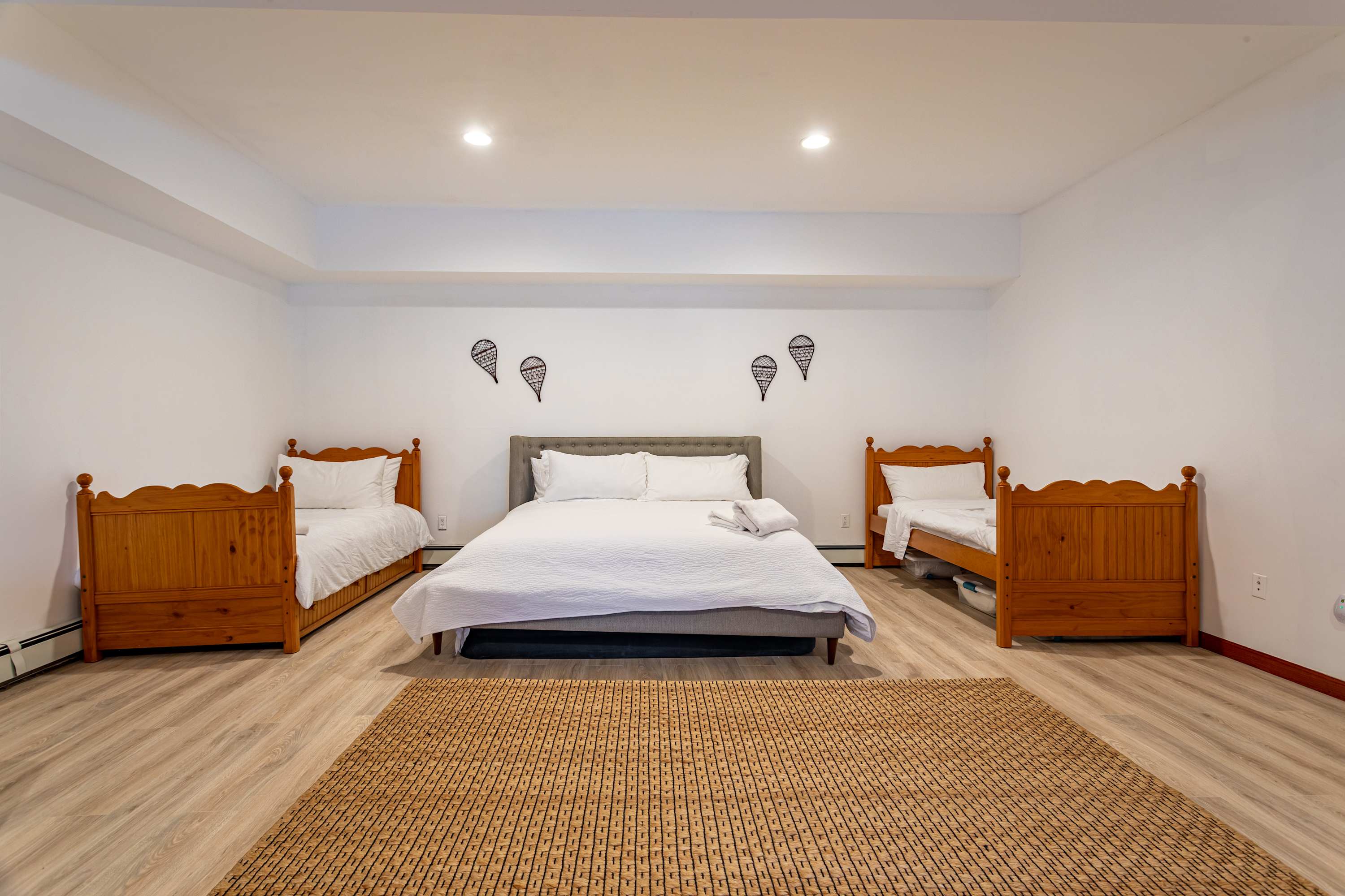 A spacious bedroom with light wood-look flooring, featuring a central queen-sized bed with a grey upholstered headboard and two wooden twin beds on either side. The room is decorated with wall art and has recessed lighting.