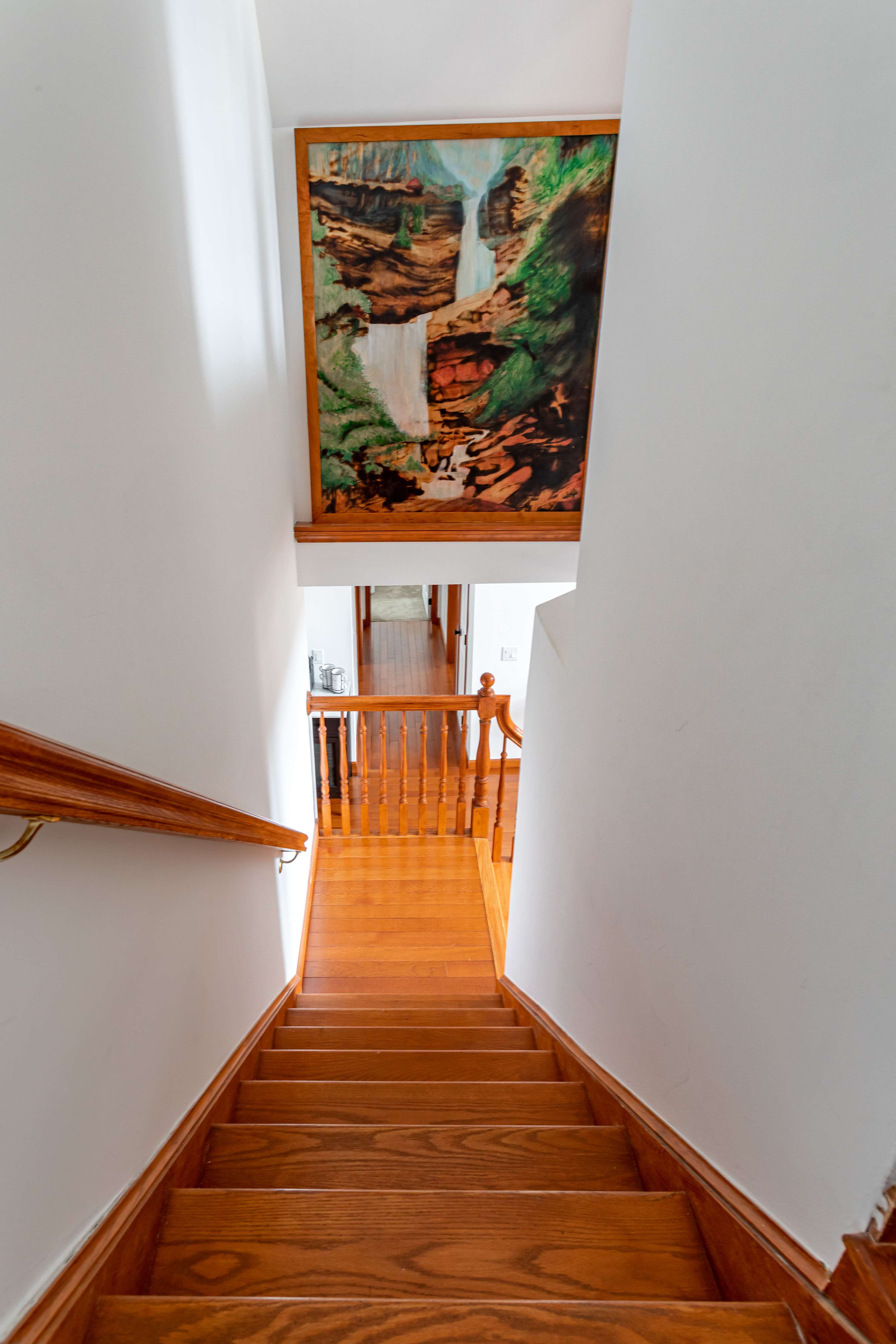 A classic wooden staircase with a matching banister leading to the lower level, with a large, vibrant framed painting of a waterfall adorning the white wall above the landing.