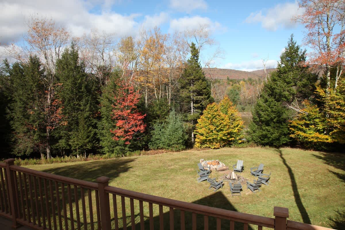 View from the wooden deck overlooking a spacious grassy yard with a fire pit area and surrounding autumn foliage. Distant mountains are visible under a partly cloudy sky, offering scenic outdoor views.