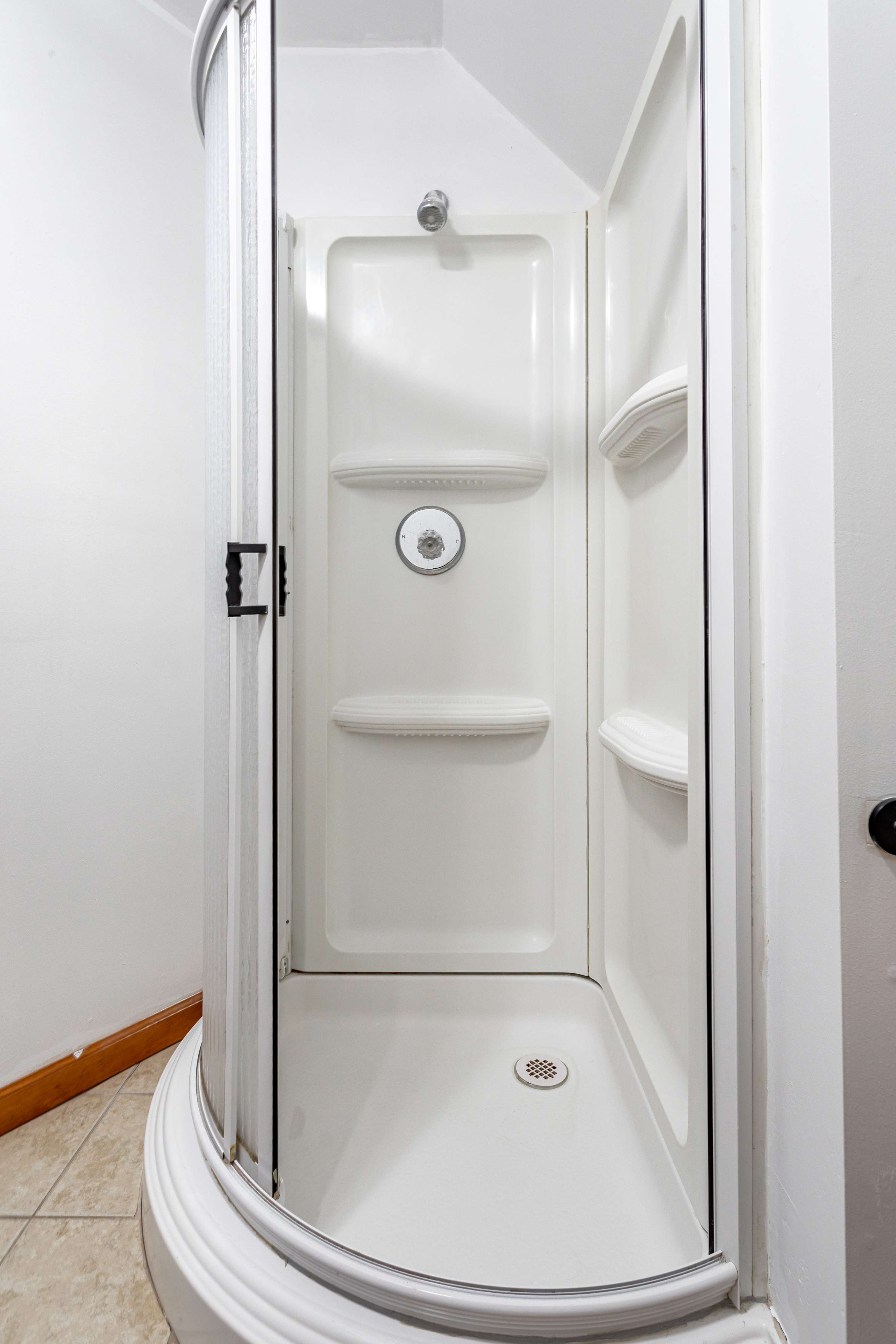 A compact, white, corner shower stall with a curved glass door, featuring built-in shelves and a standard shower head. The floor is tiled in a light brown/beige.