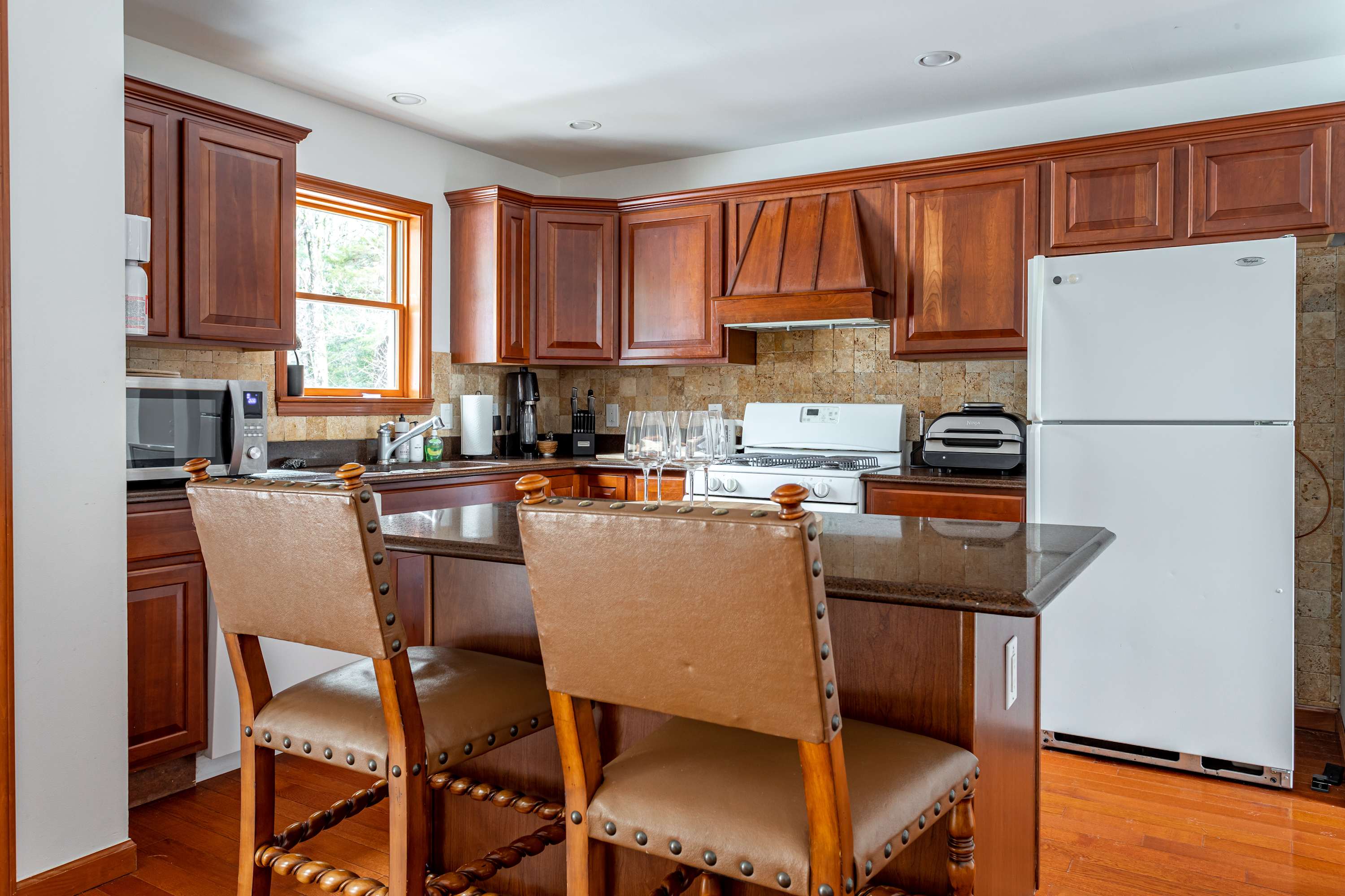 A close-up of the kitchen island with two brown leather bar stools, showcasing the dark countertops and rich wooden cabinetry. White appliances, including a refrigerator and stove, are visible in the background.