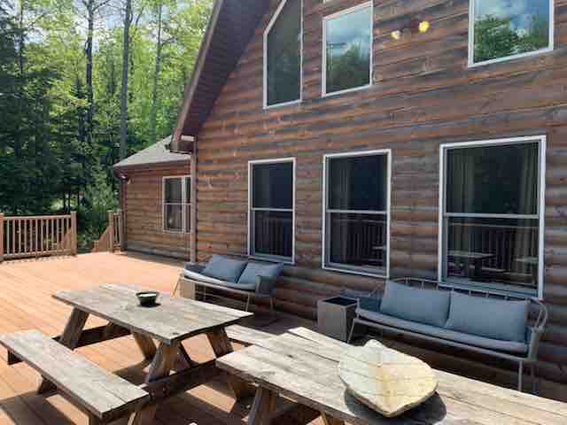The exterior of a rustic log cabin-style house with multiple windows, featuring a large wooden deck furnished with two picnic tables and built-in benches. Lush green trees surround the property.