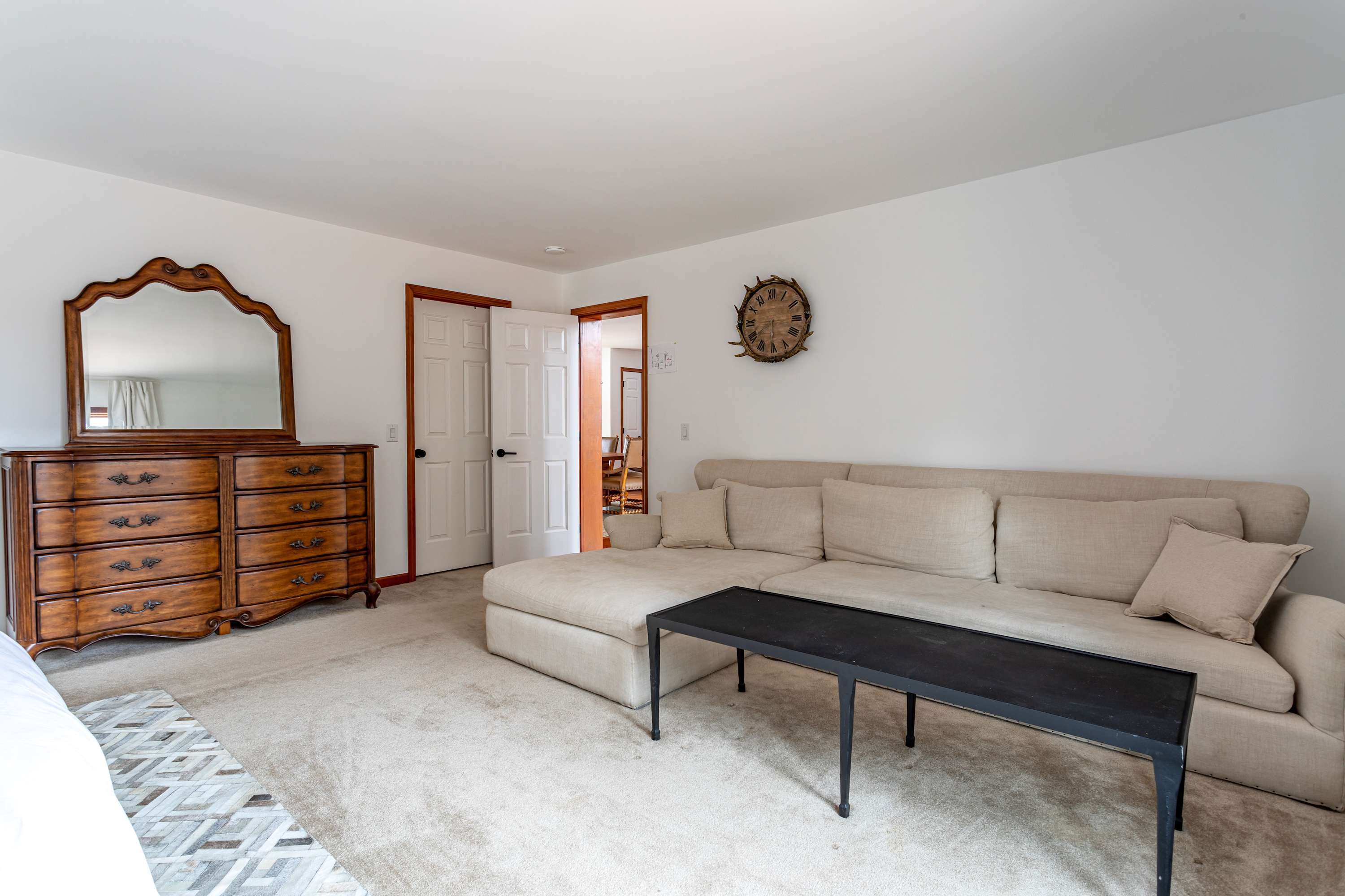 The lounge area within the master bedroom, featuring a light-colored sectional sofa, a black coffee table, a large wooden dresser with an ornate mirror, and a doorway leading to another room, all on a neutral carpet.