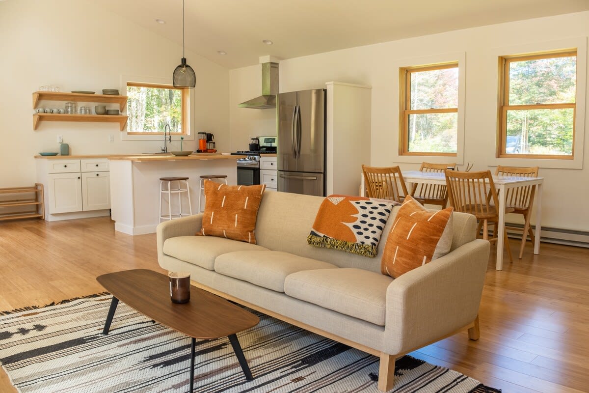 An open-concept living area with a light beige sofa, orange and black throw pillows, a dark wood coffee table on a patterned rug, and a view into the kitchen and dining area with light wood floors and white walls.